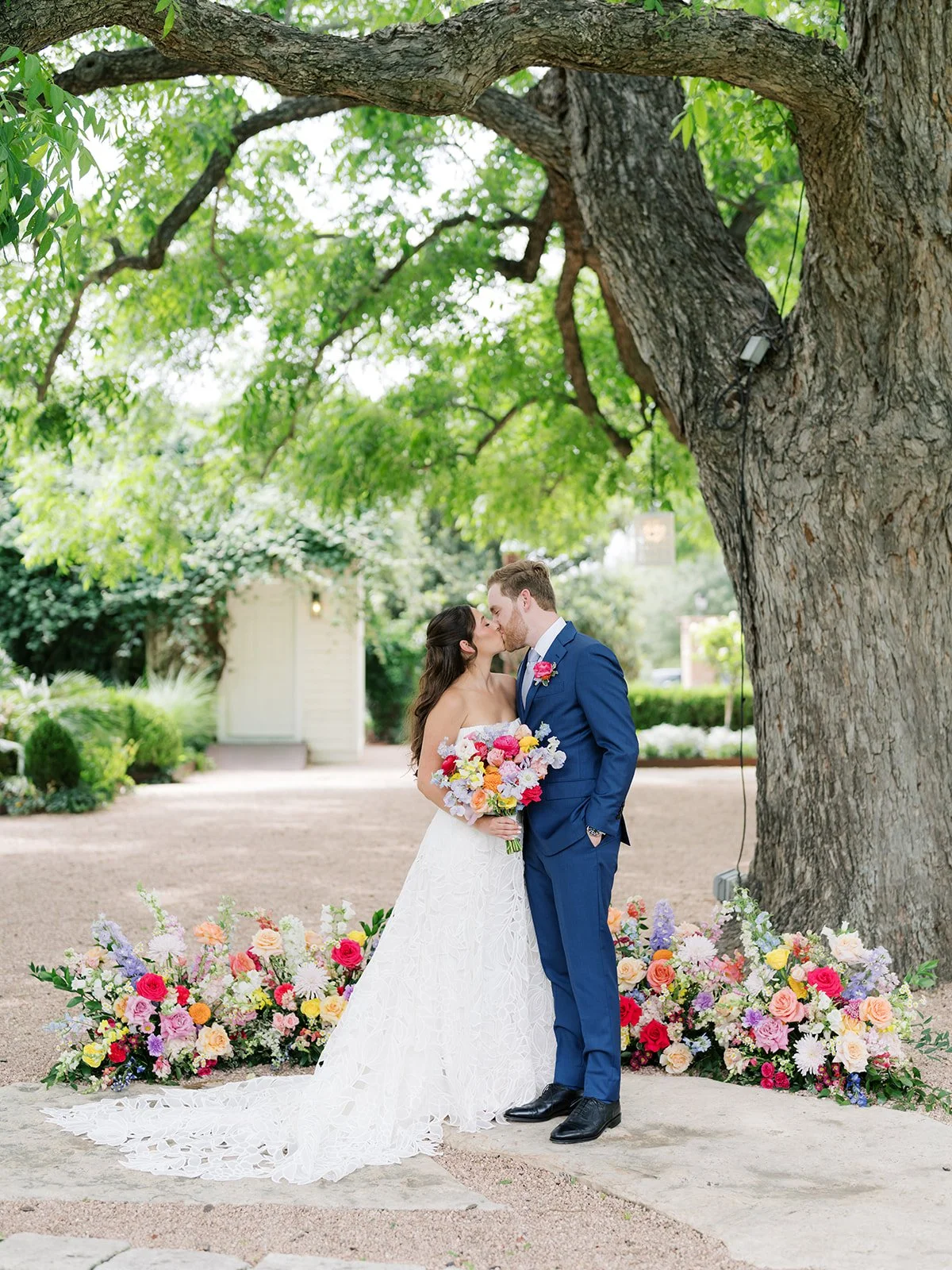 A bride and groom sharing a kiss in a garden under a large tree, surrounded by colorful flower arrangements.