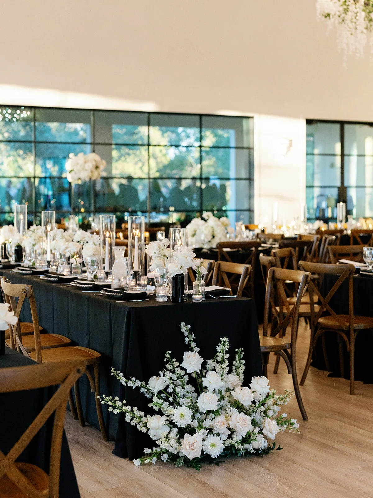 Elegant banquet hall with long black-draped tables decorated with white flowers and tall candles, large windows showing trees outside, and wooden chairs surrounding the tables.