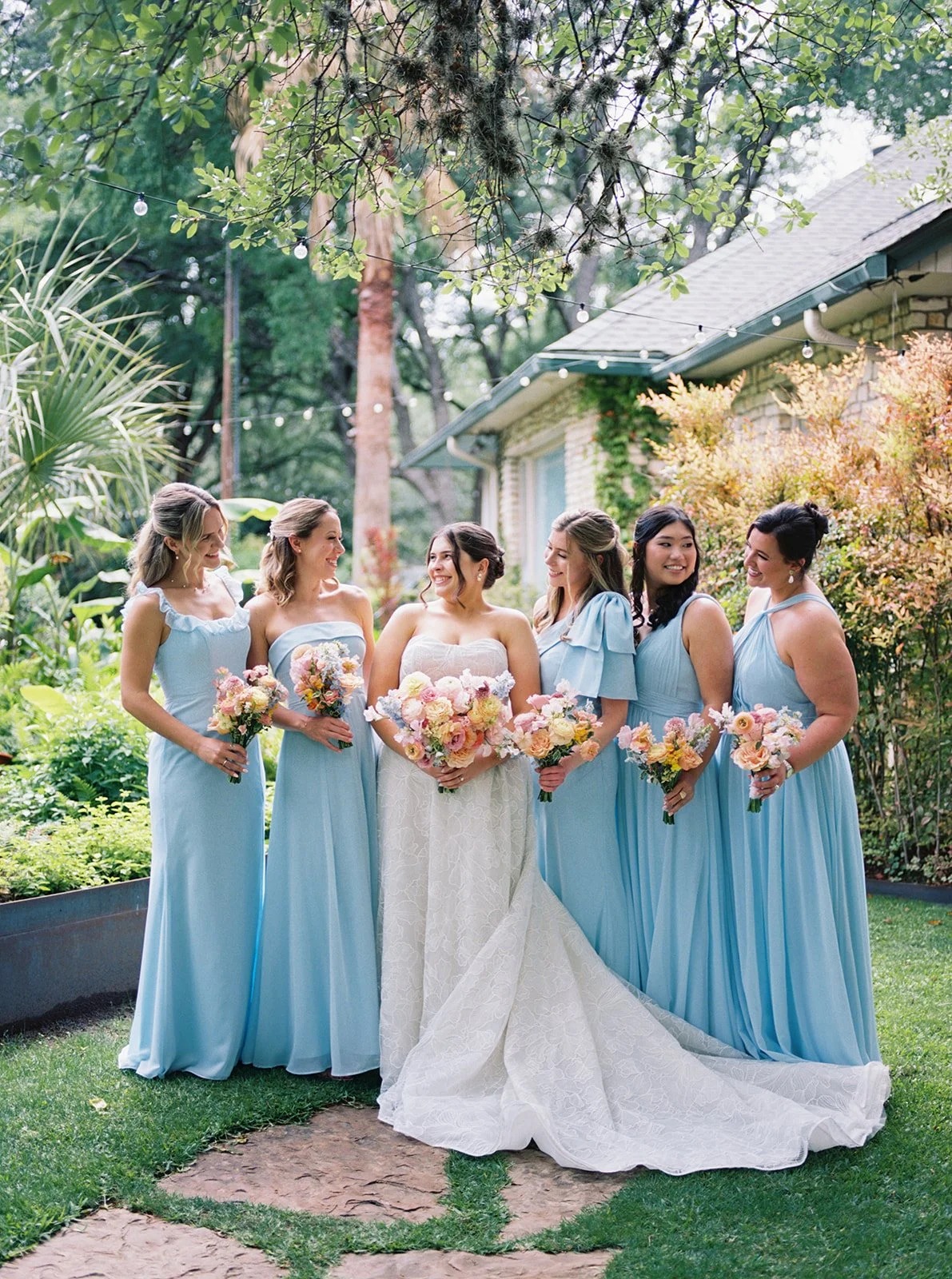 A group of six women, with one in a white wedding dress and the other five in matching light blue bridesmaid dresses, standing outdoors in a garden holding bouquets of flowers, smiling and looking at each other.