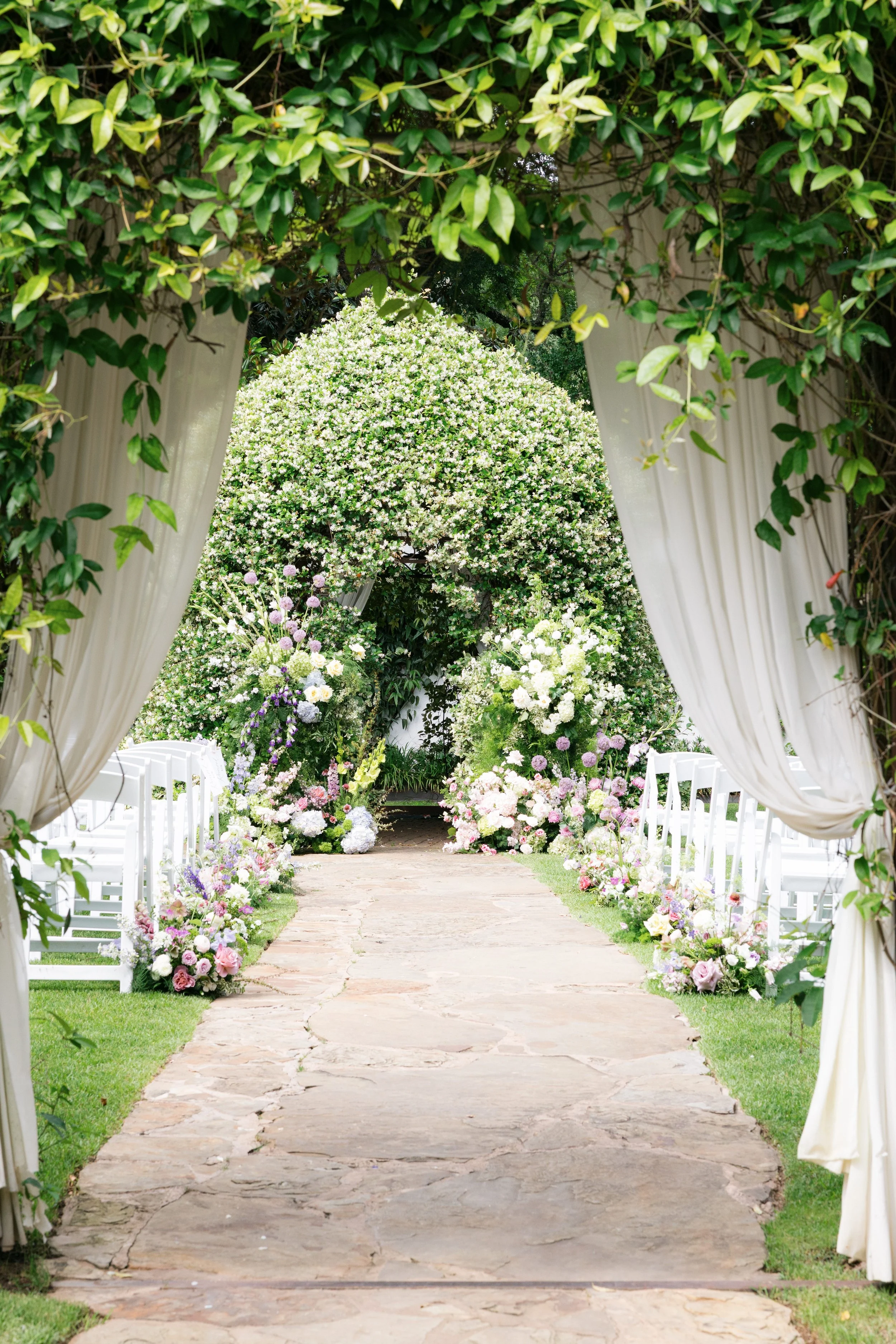 A decorated outdoor wedding aisle with white curtains and lush greenery, leading to a floral arch covered in white and pink flowers.
