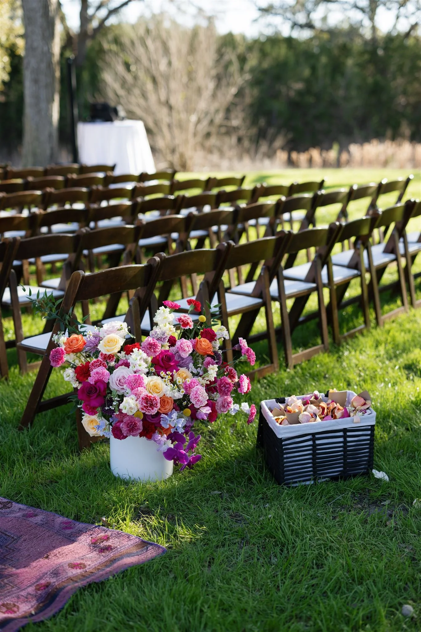 Empty outdoor wedding ceremony setup with wooden chairs, a floral arrangement in a white vase, and a basket of rose petals on a grassy lawn, with a table in the background.