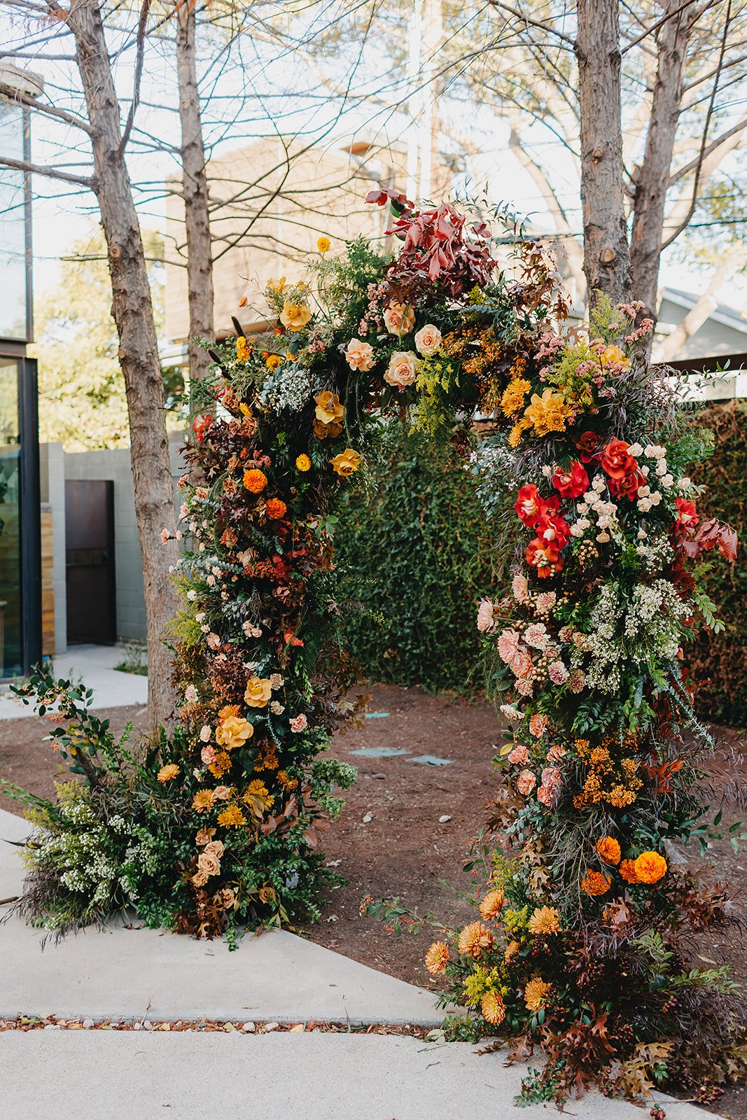 A flower arch decorated with various colorful flowers including yellow, red, pink, and orange, standing outdoors with trees and a building in the background.