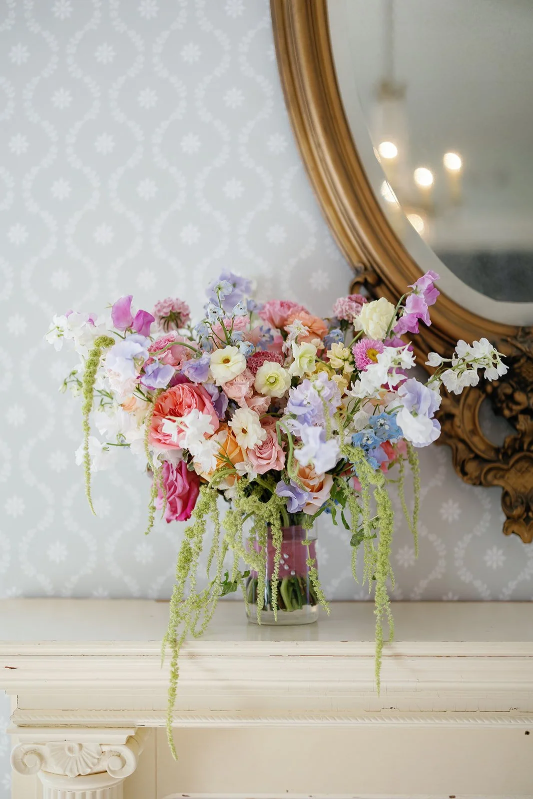 A colorful bouquet of flowers in a glass vase placed on a white mantel with a mirror and patterned wallpaper in the background.