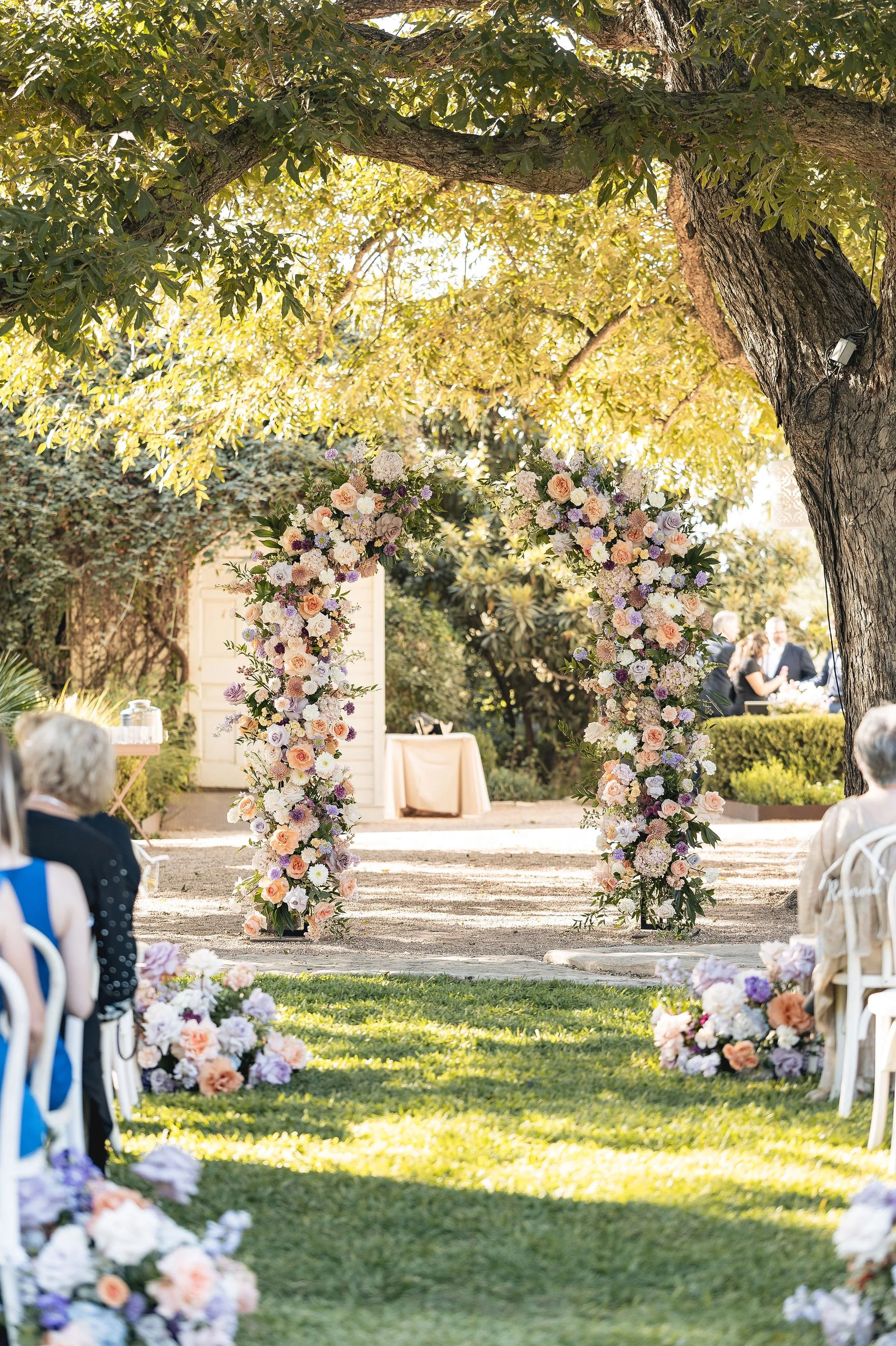 A decorated outdoor wedding ceremony area with floral arrangements on stands forming an arch under a large tree with green leaves. Guests are seated on white chairs on each side of the aisle. The background shows a sunny day with a couple, the groom 