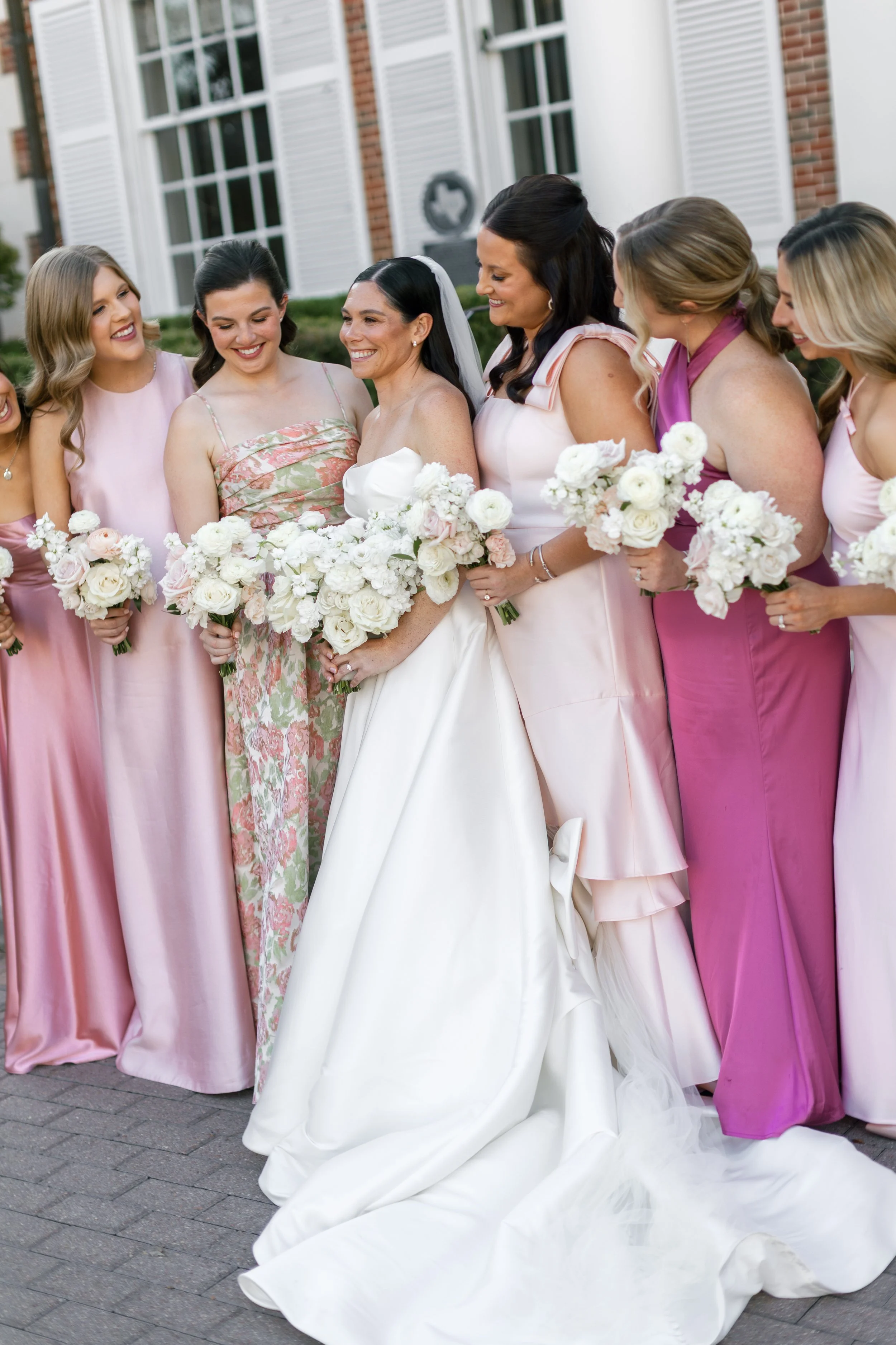 A bride in a white wedding dress surrounded by bridesmaids in various shades of pink and purple dresses, holding bouquets of white flowers.