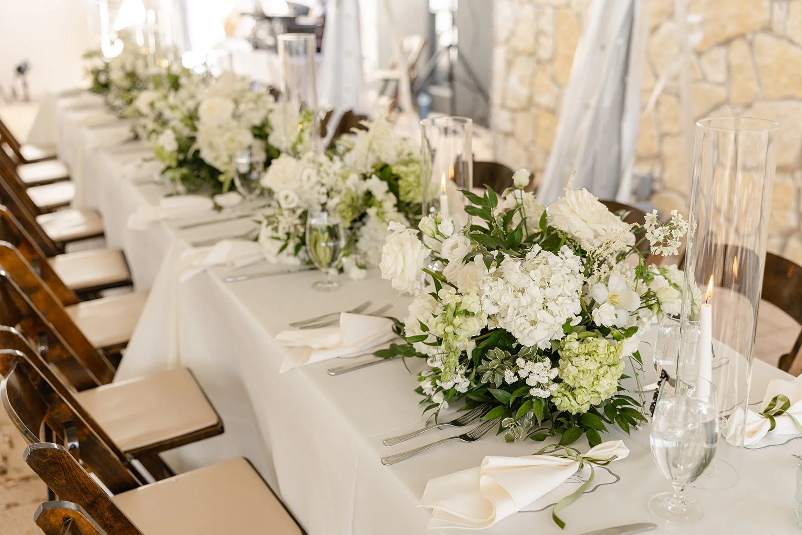 Elegant banquet table decorated with white flowers, candles, and place settings, set against a rustic stone wall.