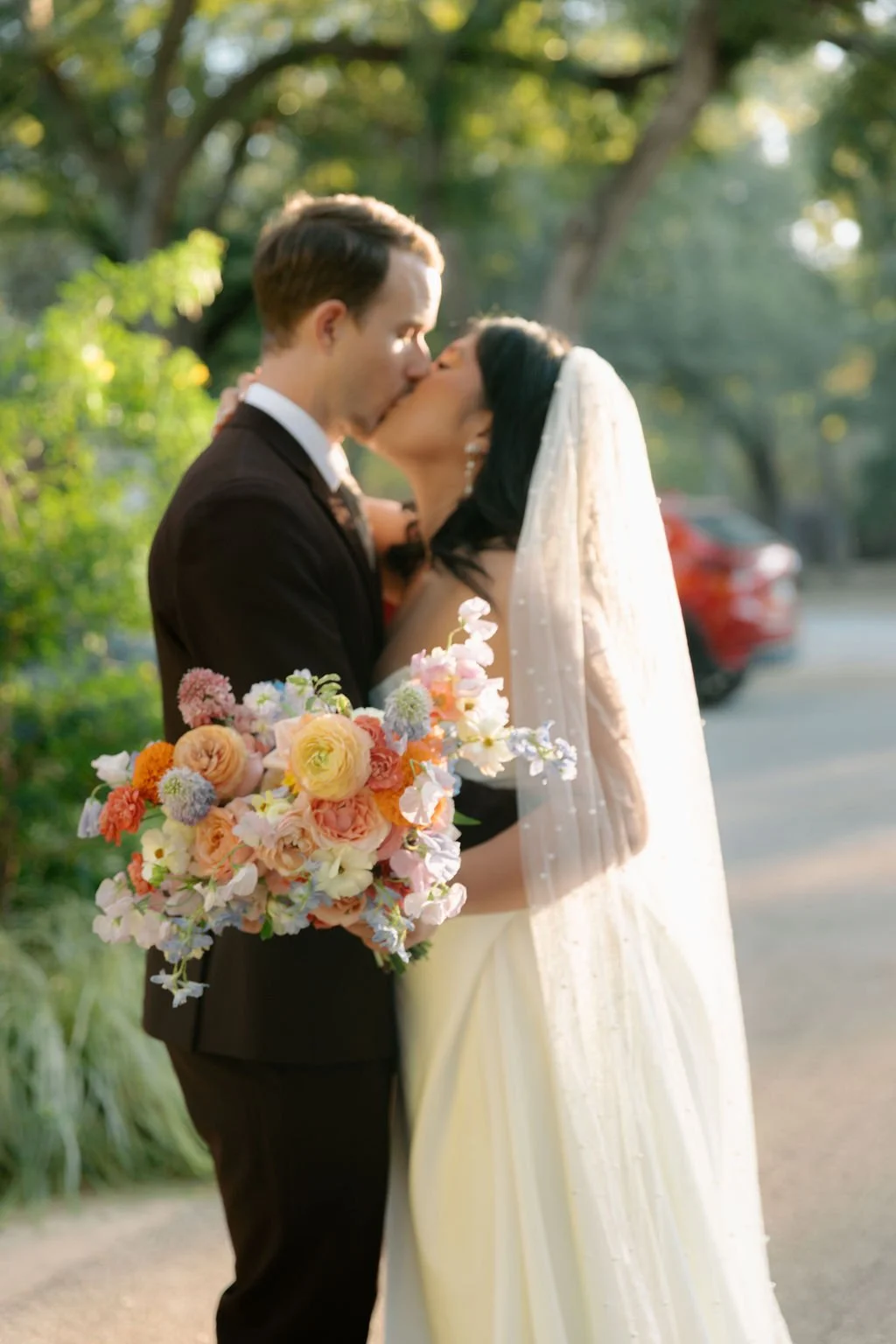 A newly married couple kissing outdoors, with the bride holding a bouquet of colorful flowers and wearing a white wedding dress and veil, while the groom is in a dark suit.