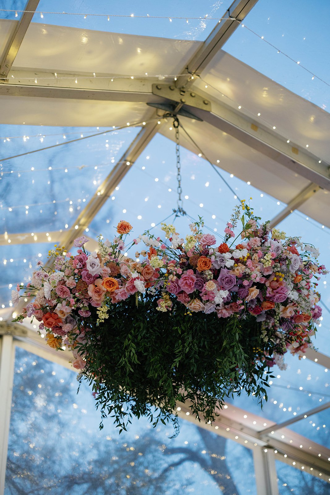 Hanging floral arrangement with pink, orange, and white flowers suspended from a ceiling with string lights, under a glass ceiling with metal framework.