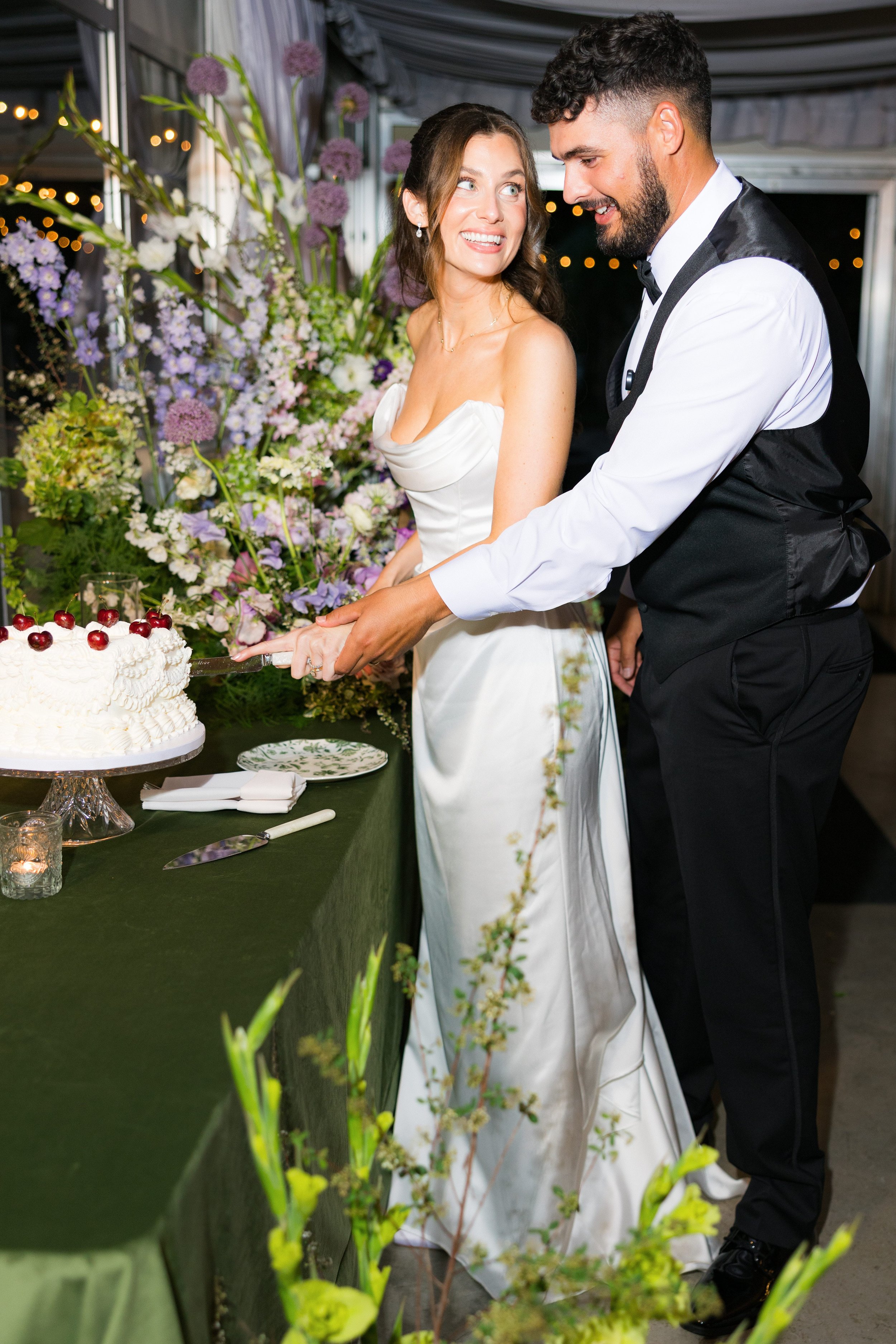 A bride and groom cutting a wedding cake together at their reception, surrounded by floral decorations and string lights.