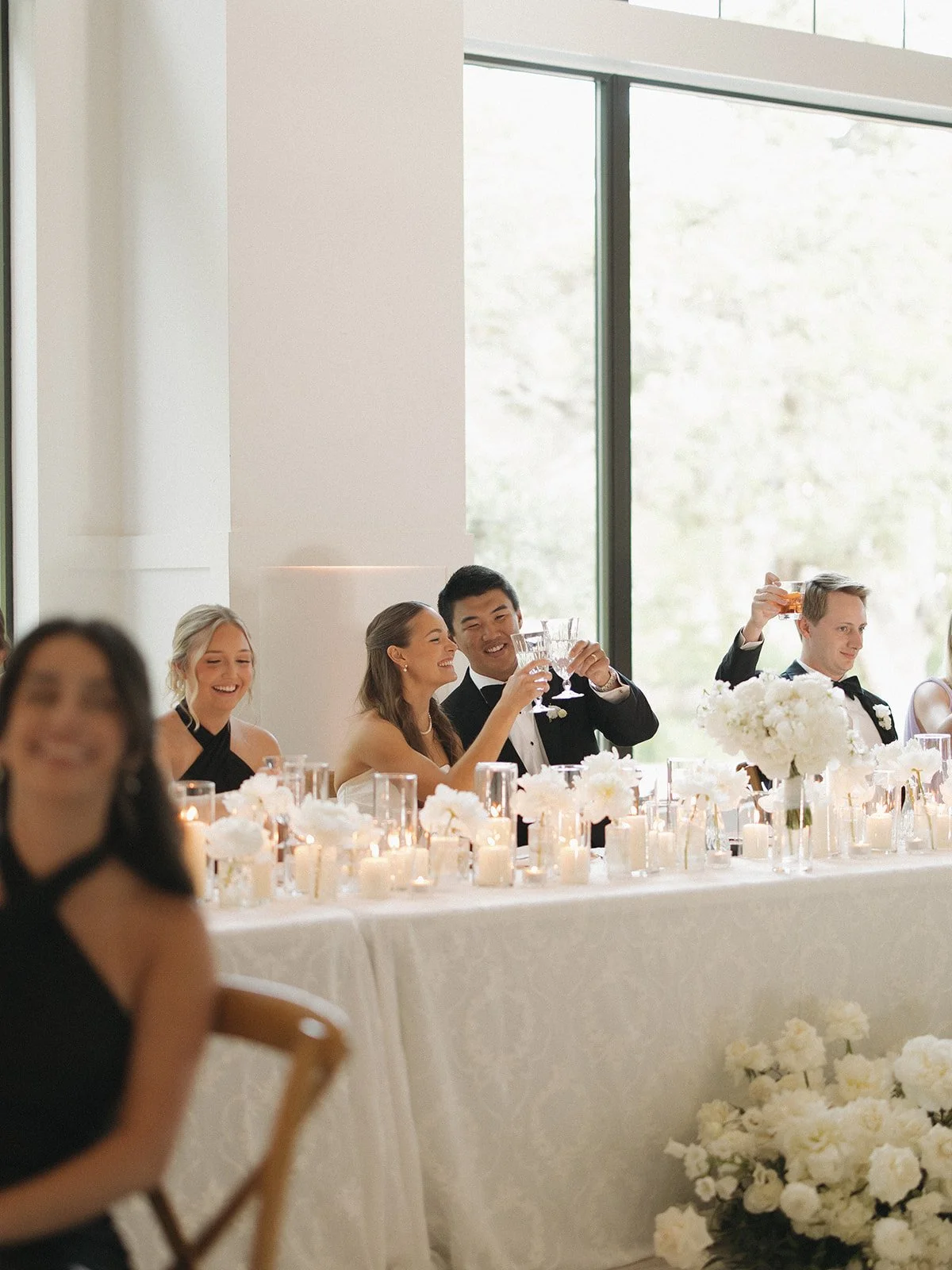 People at a wedding reception sitting at a decorated table with white flowers and candles, raising glasses for a toast, in a bright room with large windows.