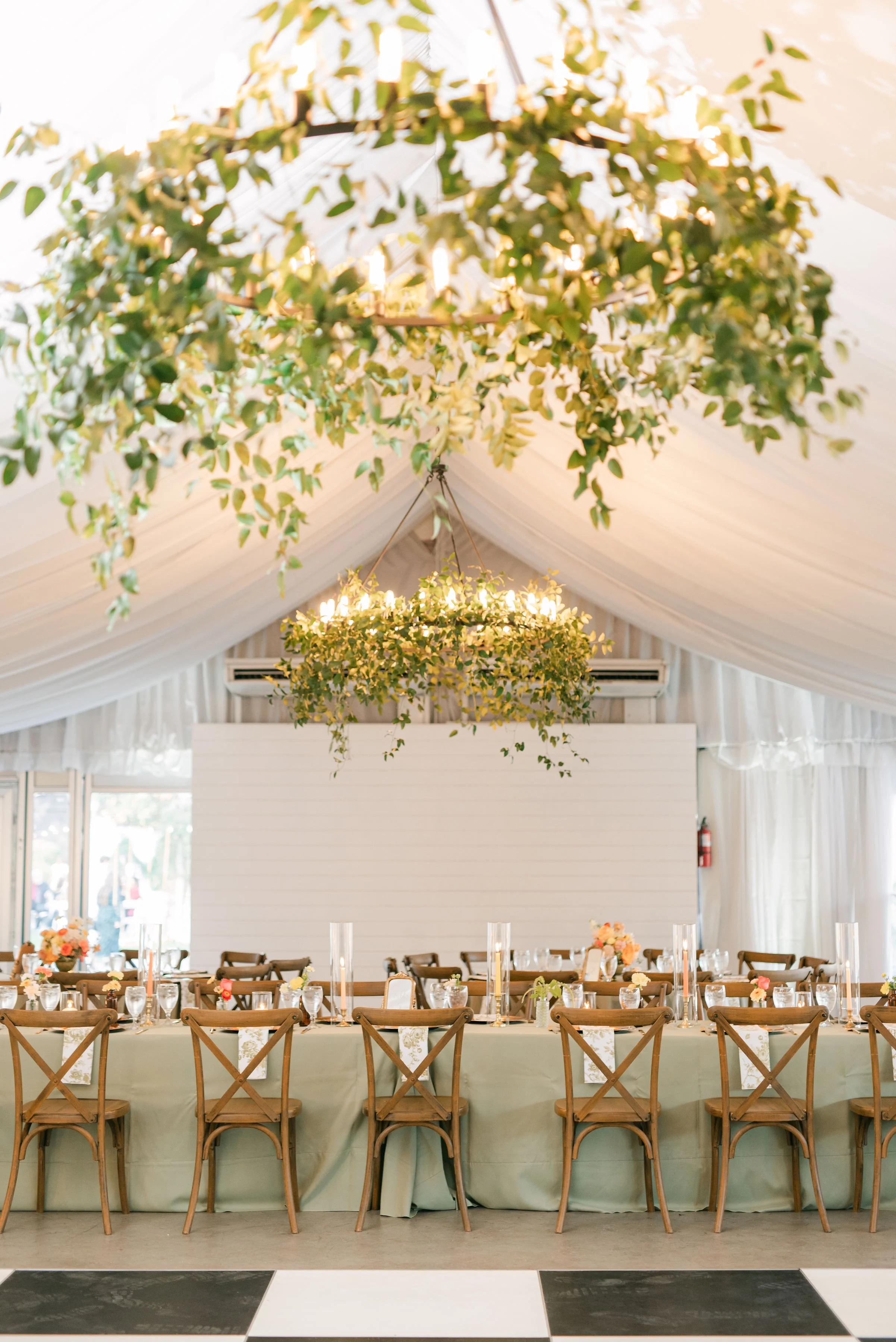 Decorated banquet table set for an event with flower centerpieces, candles, and place settings, in a room with draped ceiling and chandeliers.