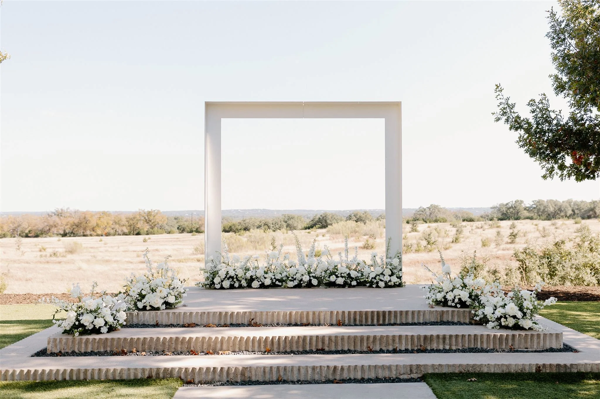 Outdoor wedding arch decorated with white flowers, set against a rural landscape with trees and open sky.