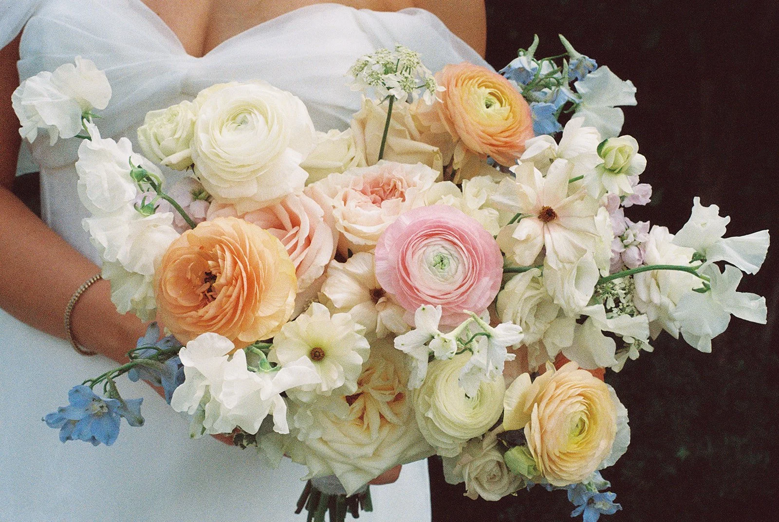 A person holding a large bouquet of various pastel-colored flowers including roses, ranunculus, and delphinium, with some person wearing a white dress and a bracelet.