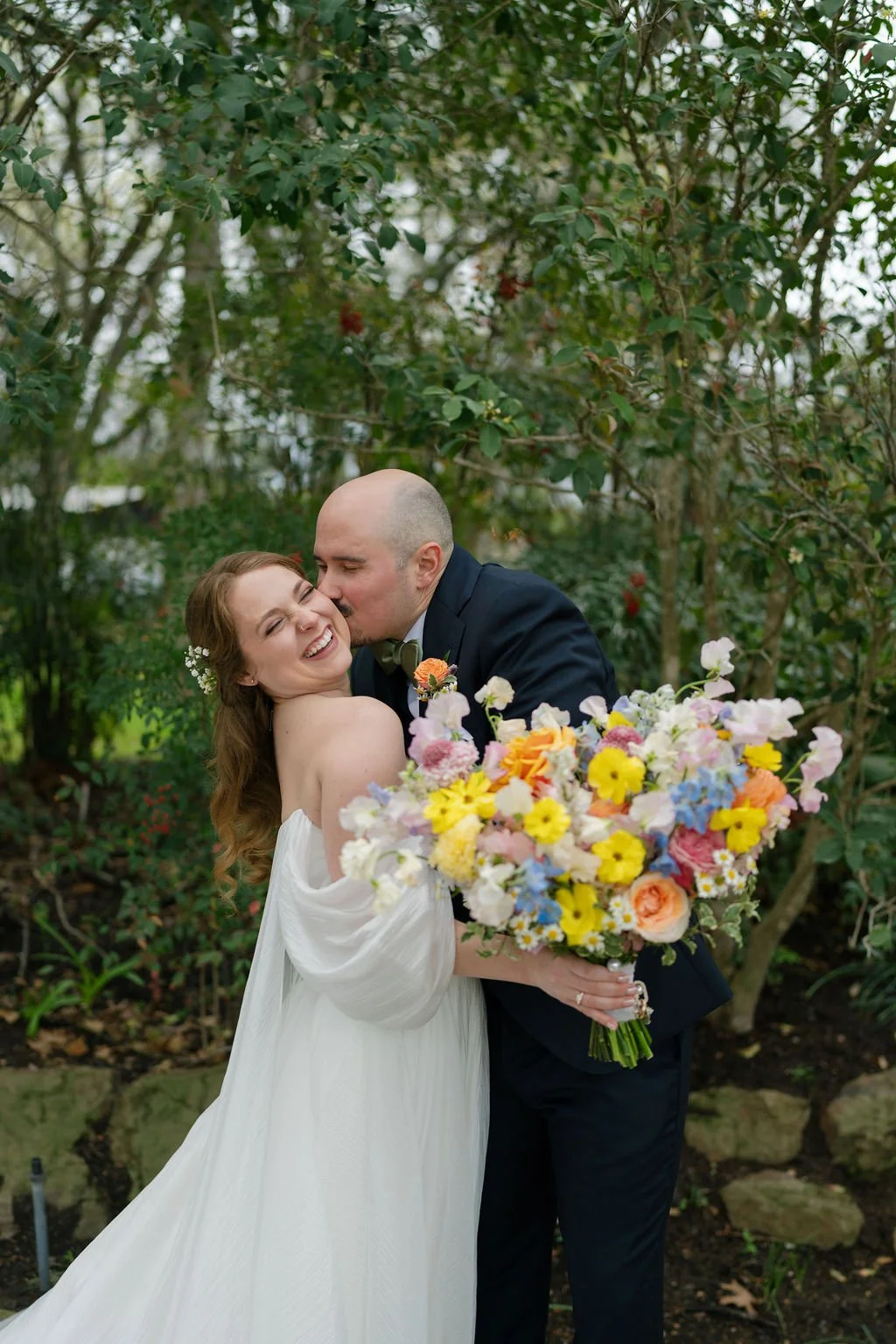 A bride and groom sharing a joyful moment outdoors, with the groom kissing the bride on her cheek. The bride is holding a large bouquet of colorful flowers. The background is filled with green foliage and trees.