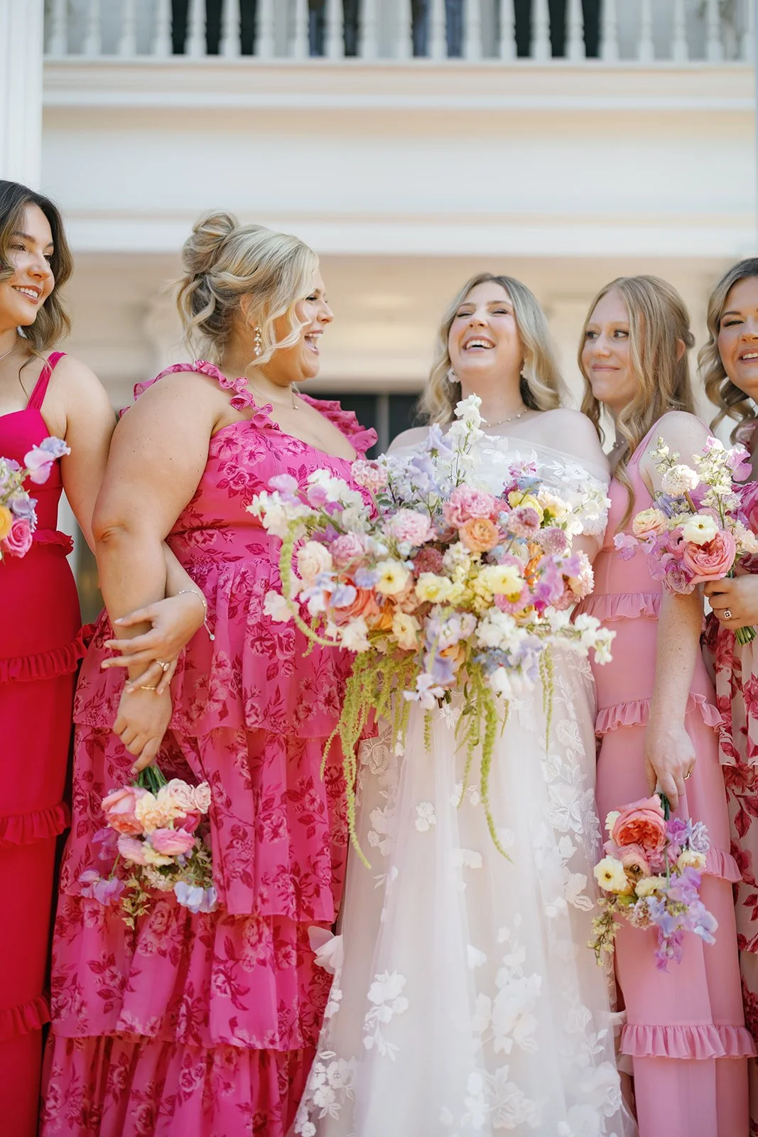 Women dressed in pink and white floral clothing, holding bouquets of flowers, smiling at a wedding celebration.