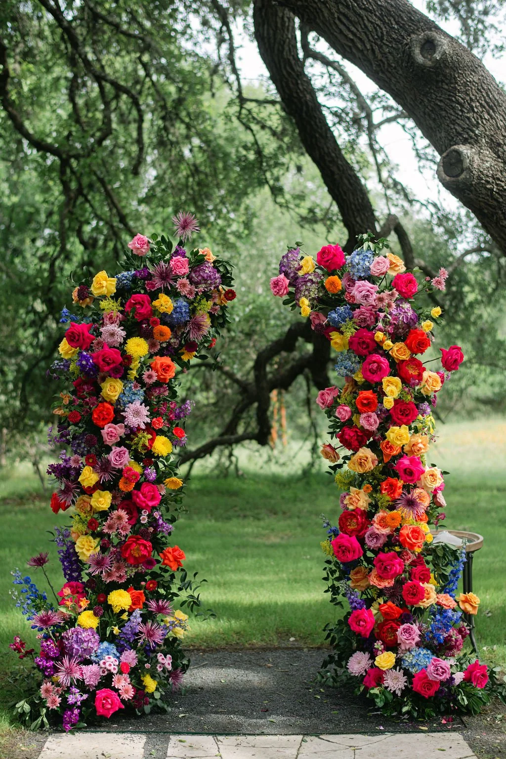 Colorful flower arch with various roses, lilies, and other flowers, set outdoors under a large tree with green foliage.