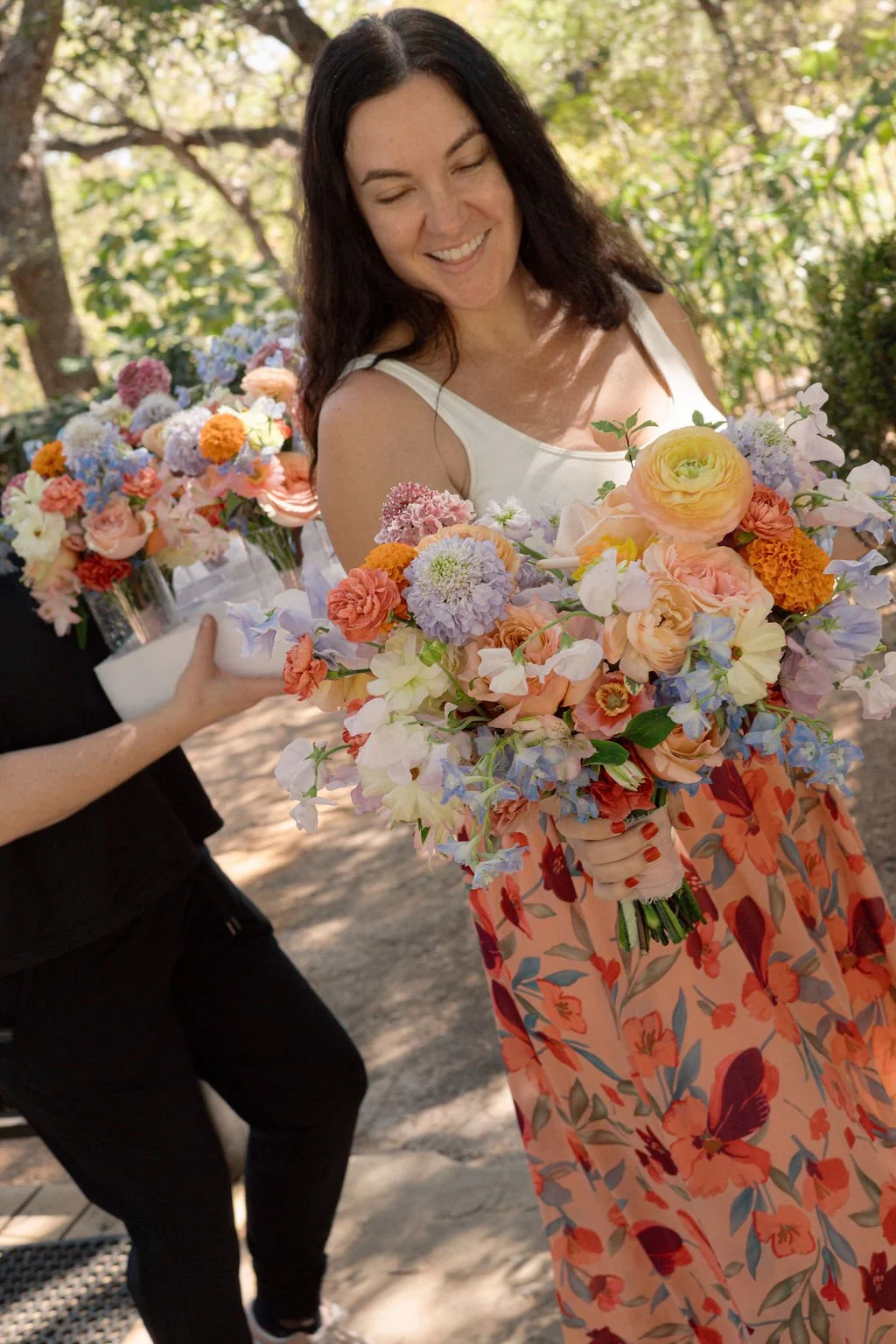 A woman holding a large bouquet of colorful flowers outdoors, smiling, with trees in the background.