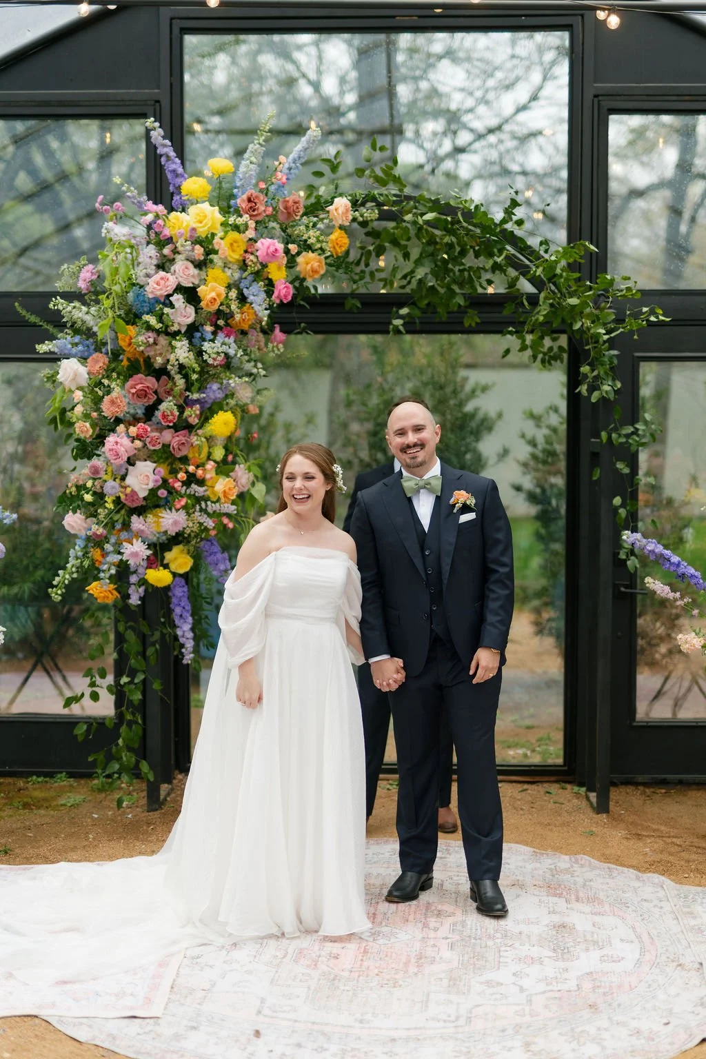 A newlywed couple holding hands and smiling at their wedding ceremony inside a glass greenhouse decorated with a large colorful floral arrangement.