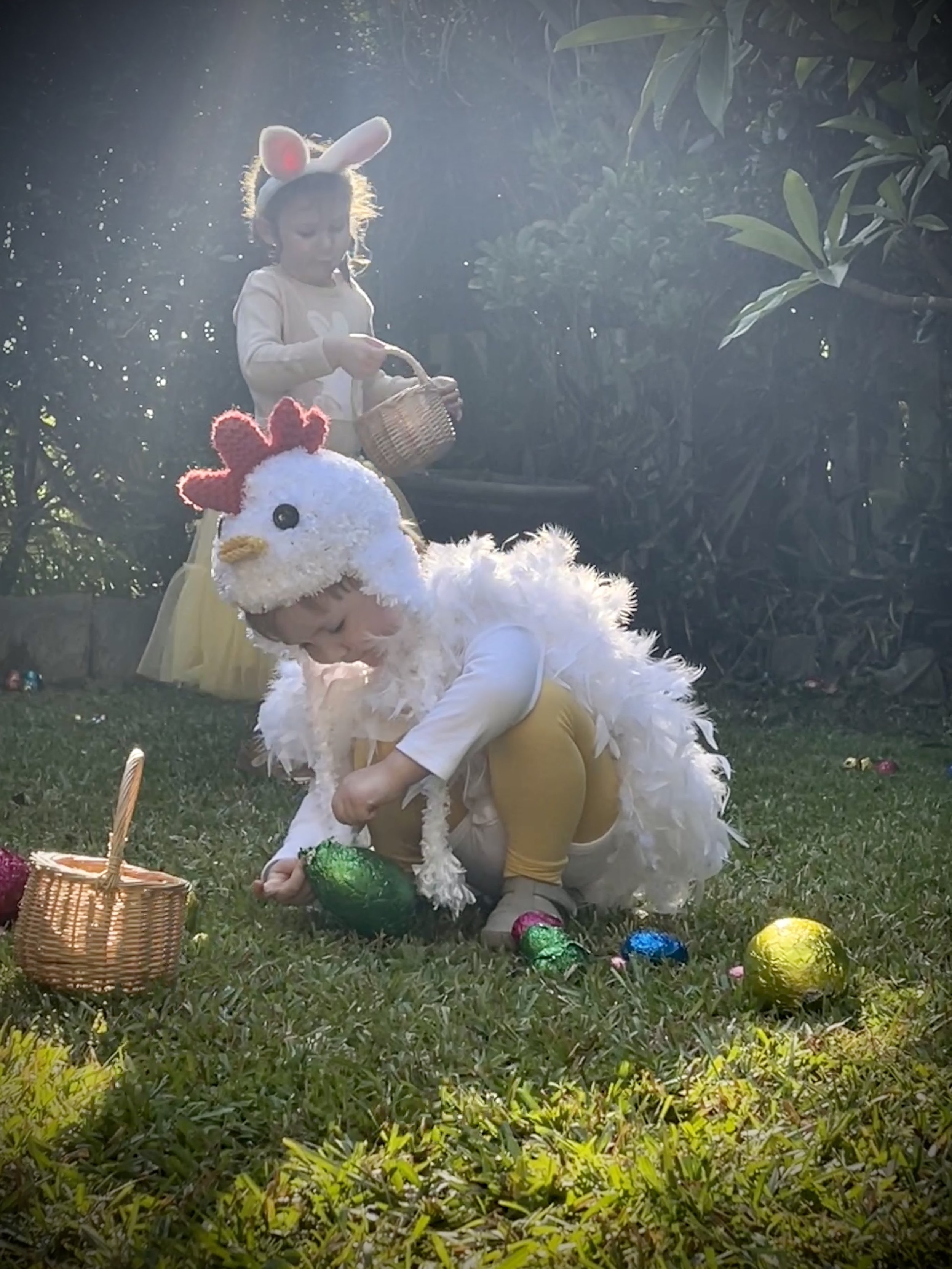 Children dressed in Easter costumes, including one in a chicken costume and another with bunny ears, engaging in an Easter egg hunt outdoors during daylight.