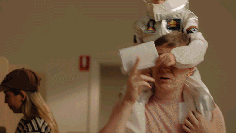 A young boy in a space suit is sitting on a woman's shoulders, covering her eyes with his arm. The woman is holding a piece of paper and a card, and appears to be emotional. A girl with long hair and a cap is walking in the background.