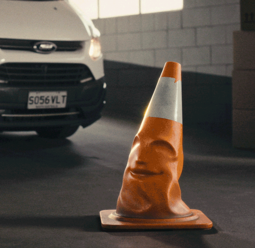 A traffic cone with an unusual distorted face illustration on it, placed on a parking lot near a white car.