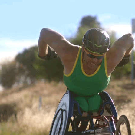 Athletic man in a green and yellow sports outfit and helmet competing in a wheelchair race outdoors on a sunny day.