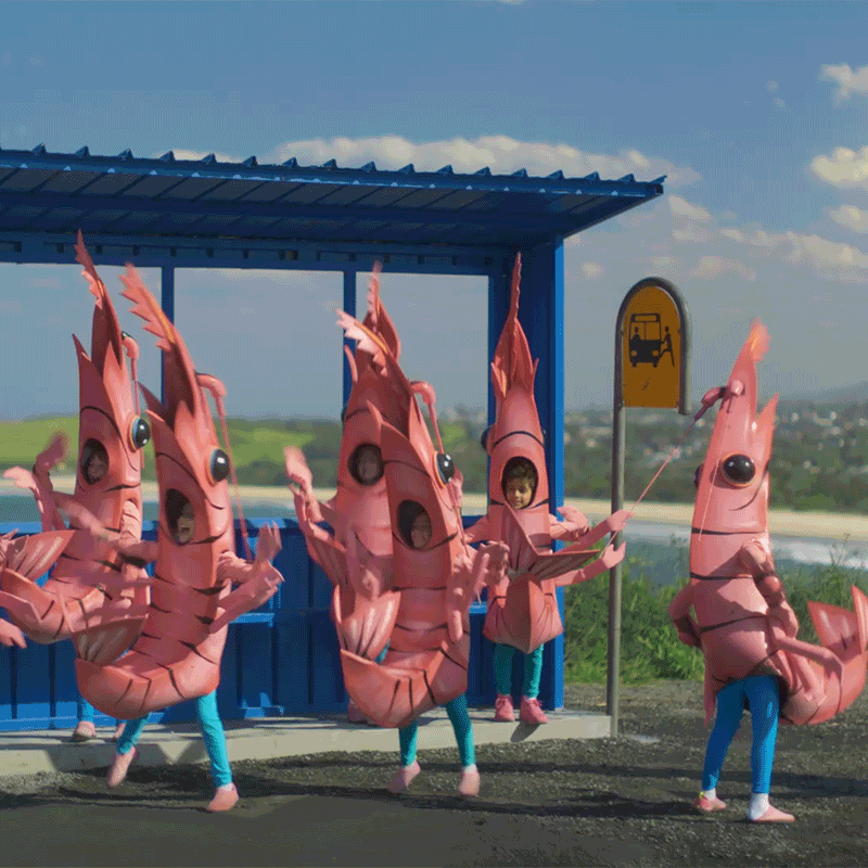 People dressed in pink lobster costumes at a bus stop on a sidewalk, with a sky and distant landscape in the background.