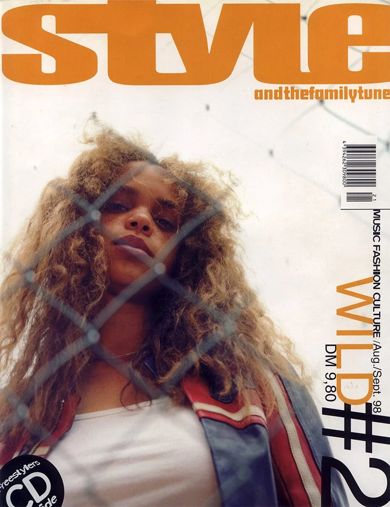Cover of a magazine featuring a woman with curly hair behind a chain-link fence.