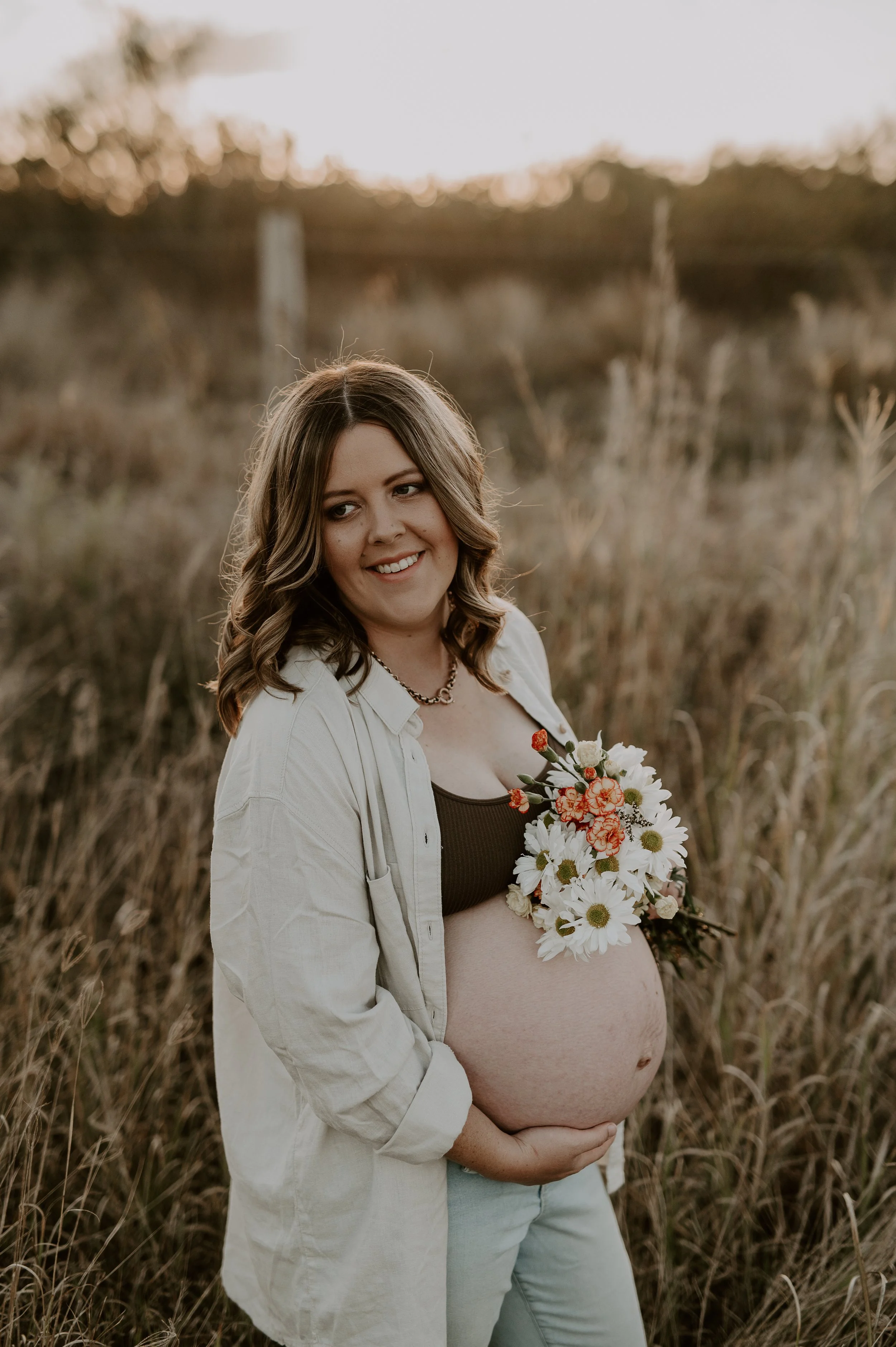 A pregnant woman standing in a field of tall grass at sunset, smiling and holding a bouquet of white and orange flowers.