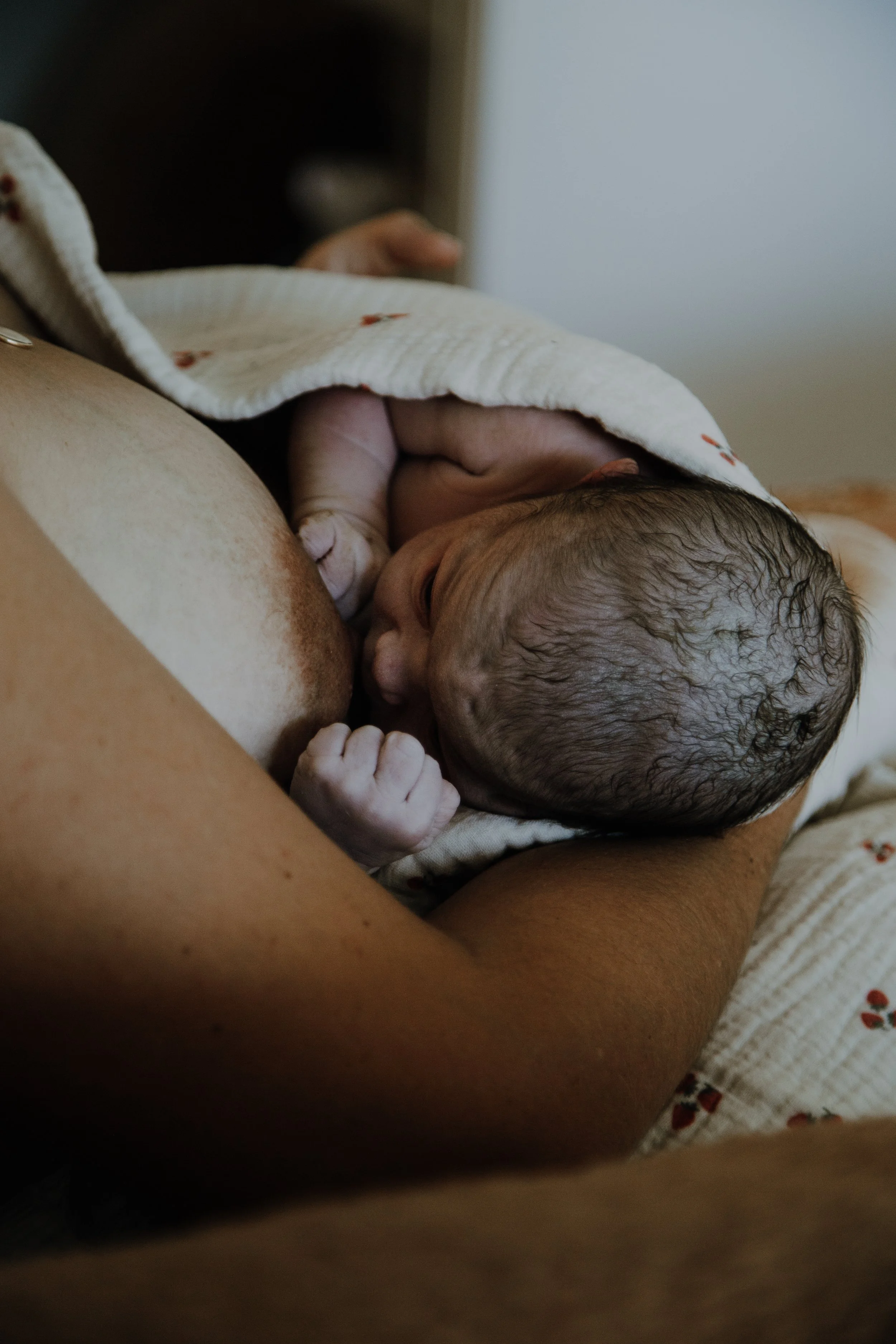 A newborn baby breastfeeding from a mother's breast, wrapped in a white blanket with small red patterns.