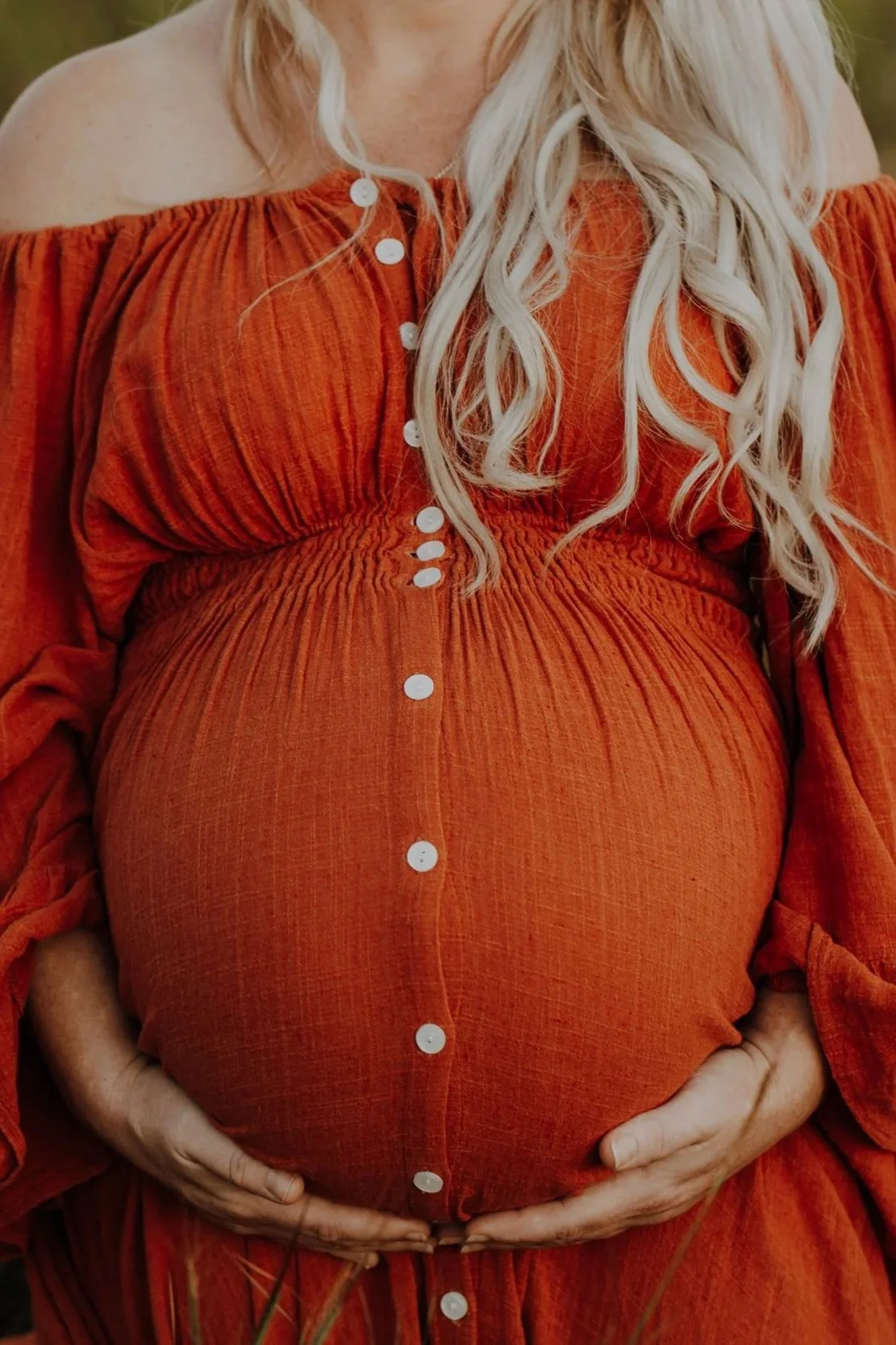 A pregnant woman wearing an orange off-shoulder dress with buttons, holding her belly with both hands.