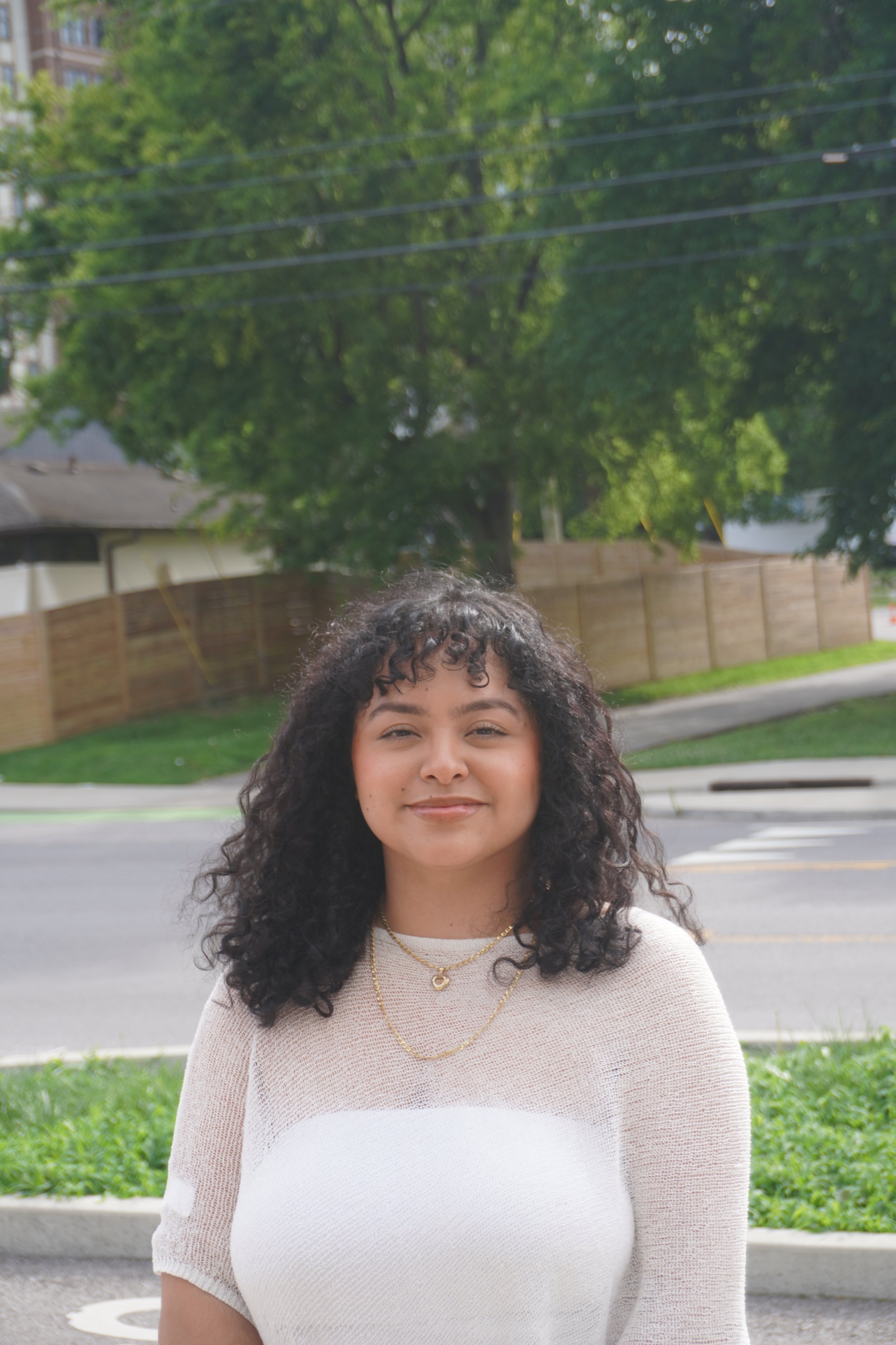 A woman with curly reddish-brown hair and light skin, wearing a gold heart-shaped necklace, smiling gently, standing against a pink brick wall.