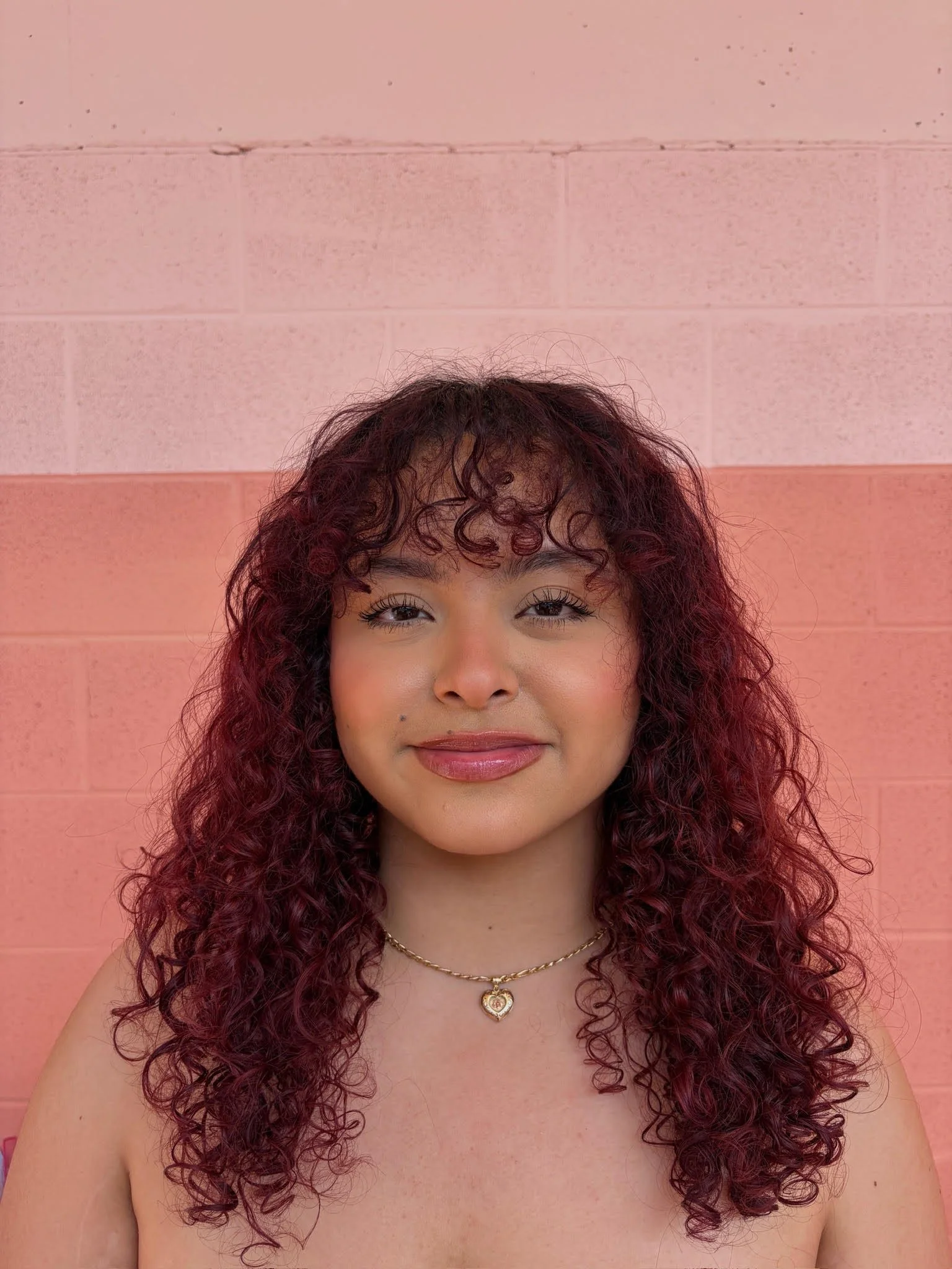 A woman with curly reddish-brown hair and light skin, wearing a gold heart-shaped necklace, smiling gently, standing against a pink brick wall.