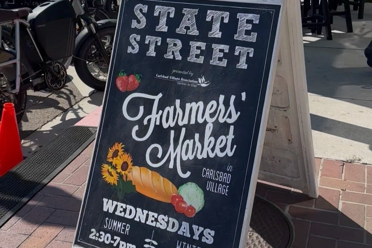 Signboard for State Street Farmers Market in Carlsbad Village promoting Wednesdays from 2:30 to 7 pm, featuring illustrations of strawberries, sunflower, carrot, and berries, with some parked bicycles in the background.