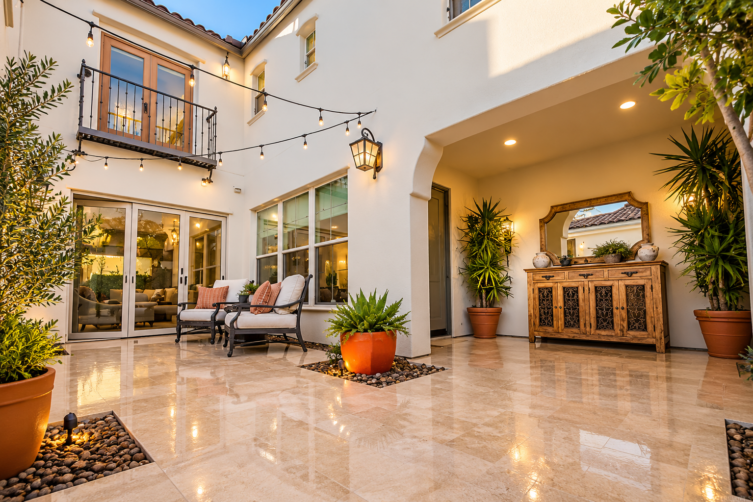 A cozy outdoor patio with potted plants, a wooden sideboard with decorative items and a mirror, seating area with cushions, string lights, and a lantern-style wall light, in a house with large glass sliding doors and beige tiled flooring.