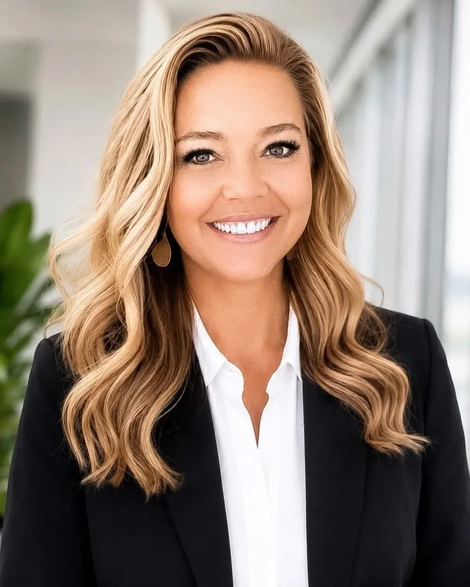 A smiling woman with wavy blonde hair wearing a black blazer and white shirt, standing indoors with a blurred background and a green plant.