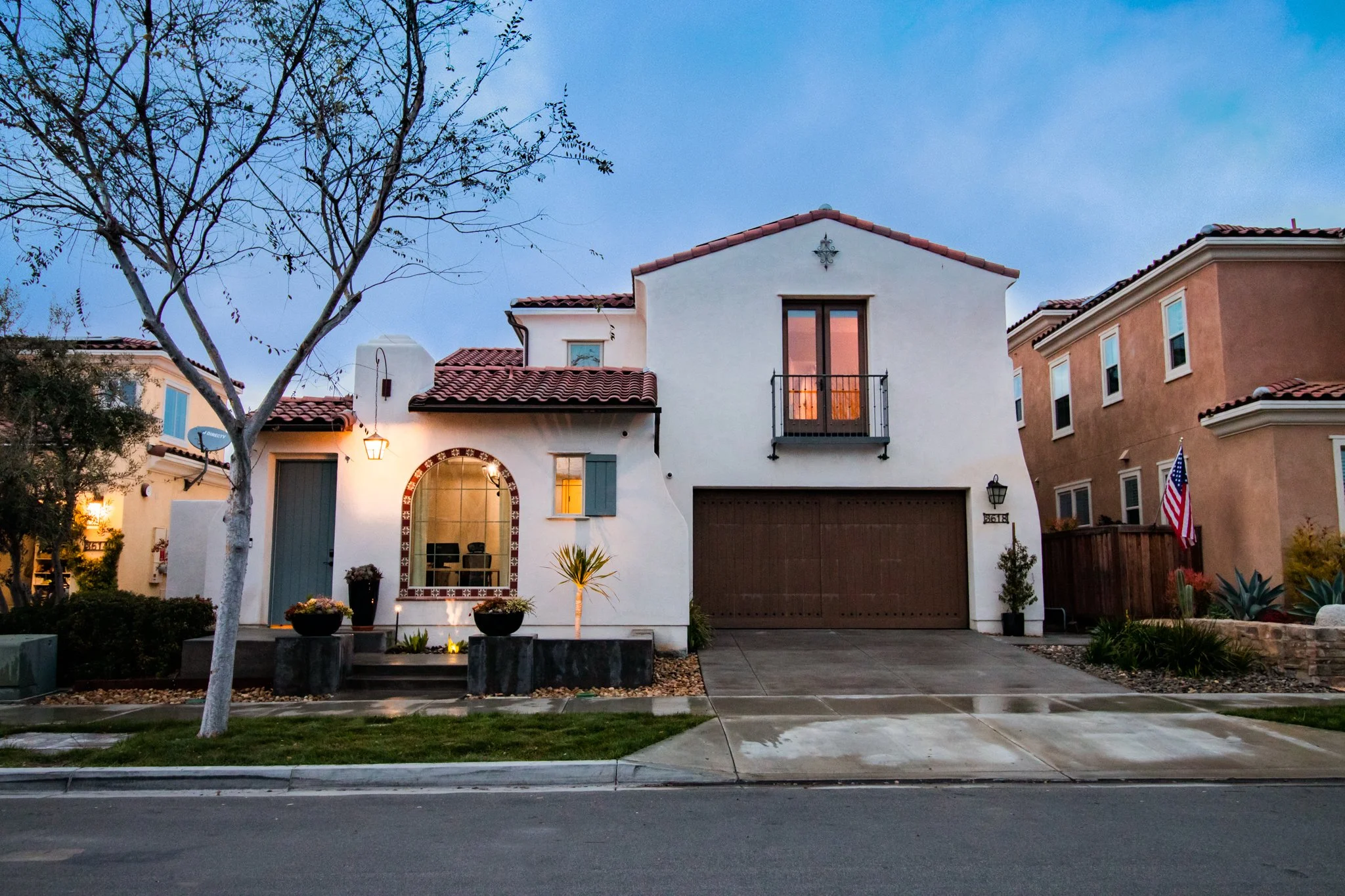 Front view of a residential house at dusk with a white facade, red-tiled roof, glass doors, small balcony, and front yard with plants, sidewalk, and street in the foreground.