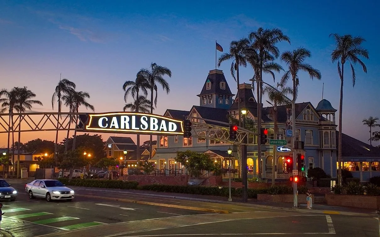 The image shows the Carlsbad village sign in front of a historic-style building with palm trees, traffic lights, cars on the street, and a sunset sky.