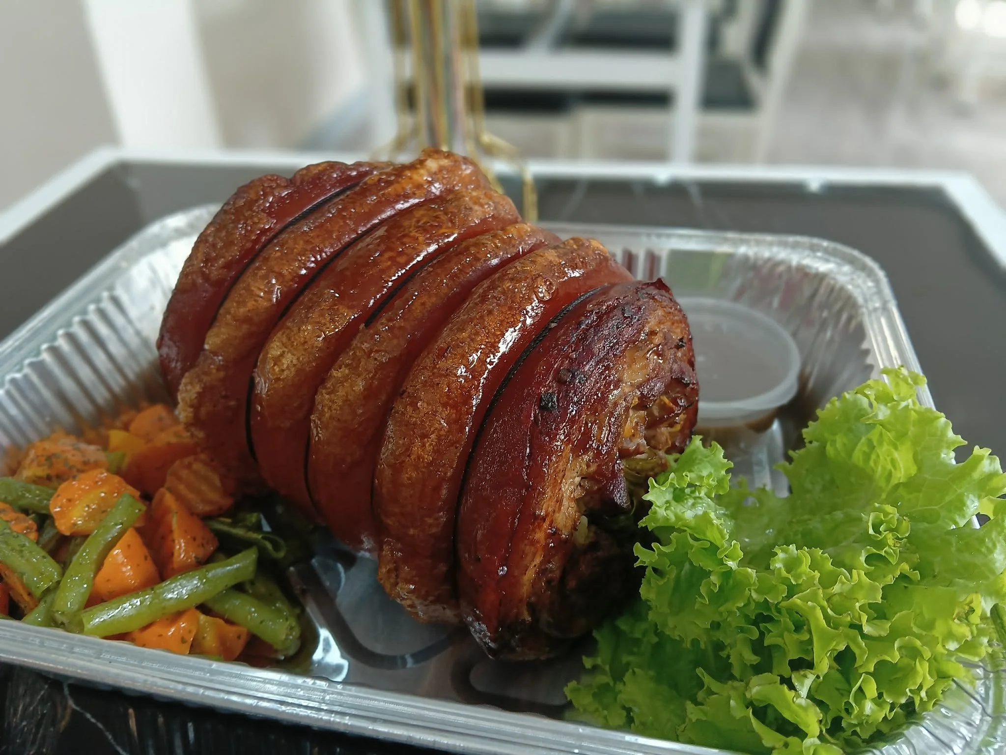 A loaded aluminum takeout container with sliced meatloaf topped with ketchup, surrounded by green lettuce, roasted carrots, and green beans.