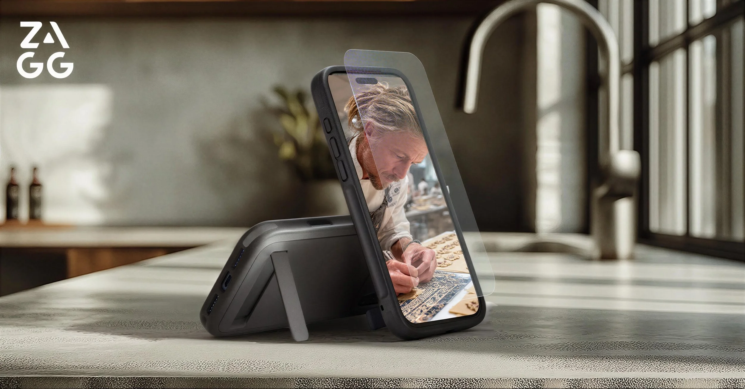 A man working on an intricate art piece at a table, with a smartphone propped up in front of him, displaying his image. The setting appears to be a modern indoor space with natural light.