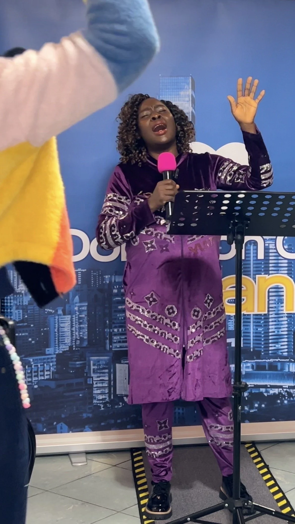 A woman singing into a pink microphone with her eyes closed and one hand raised, standing on a small skateboard, in front of a cityscape background.