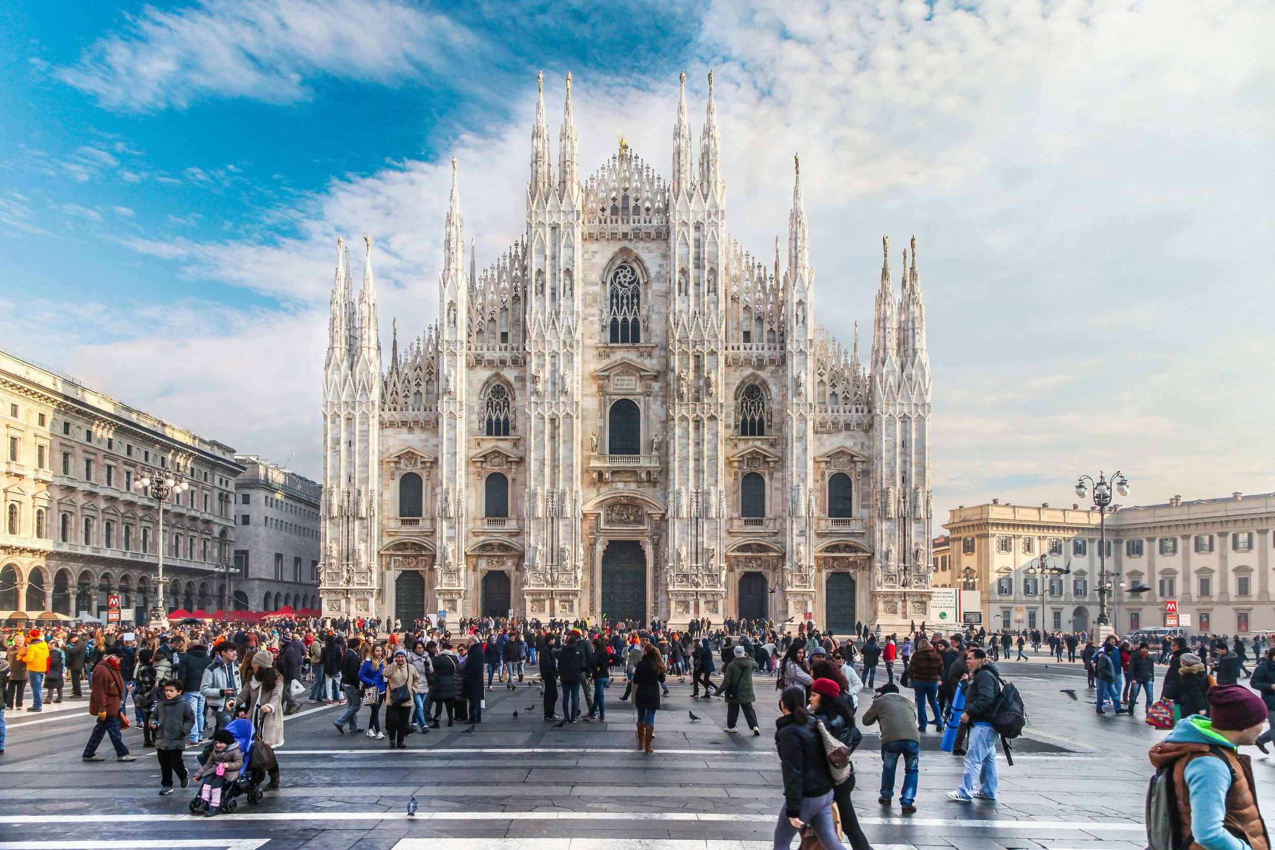 The Milan Cathedral, a large white Gothic church with multiple spires, is in the background. The cathedral is highly detailed and ornate. In the foreground, many people are walking and gathering in a busy city square under partly cloudy skies.