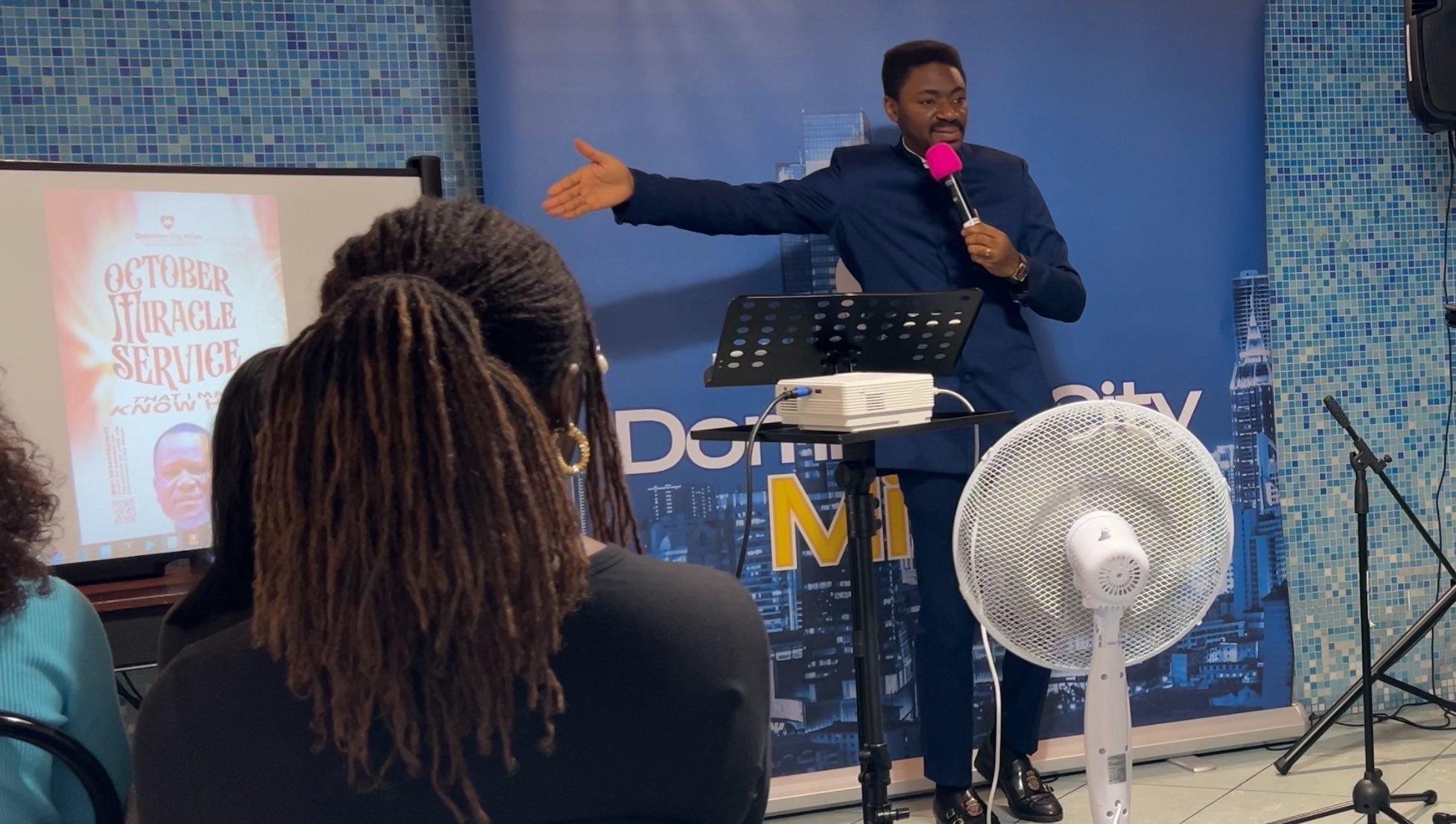 A man in a dark suit presenting to an audience in front of a blue cityscape background with a large fan and microphone. A screen displays a poster titled "October Miracle Service" with a picture of a man.