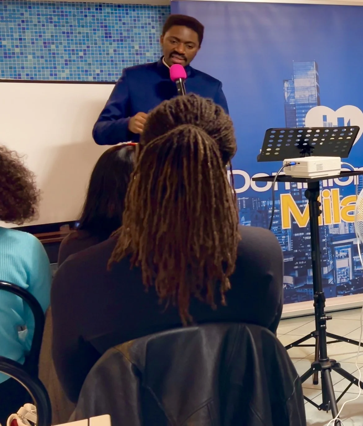 A man in a blue suit giving a presentation at a conference, speaking into a microphone with a pink cover, in front of a blue cityscape backdrop and an audience of women with various hairstyles.