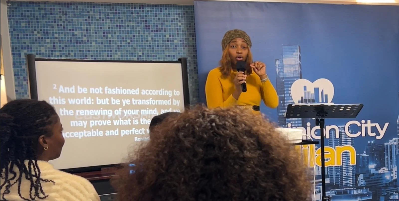 A woman in a yellow outfit and headwrap is giving a presentation, holding a microphone, in front of a backdrop with a cityscape and the words 'Urban City' in part. There is a screen displaying a quote and a music stand with papers.
