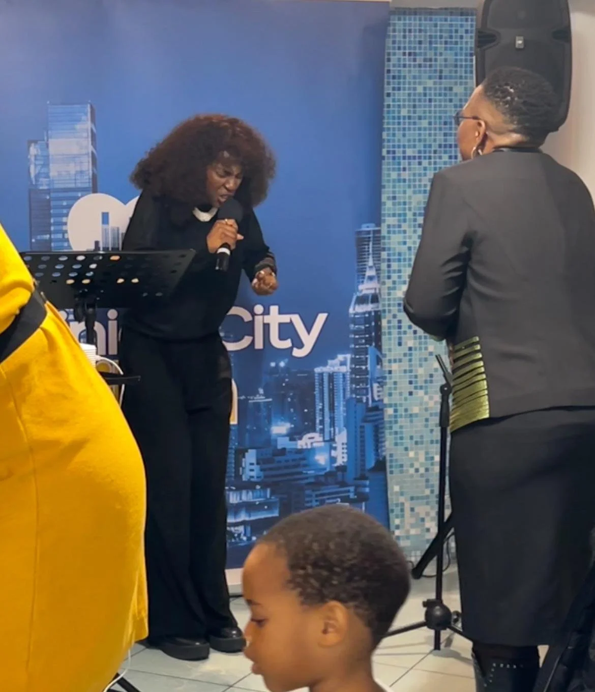 A woman in a black outfit singing passionately into a microphone during a live performance at an event with a city skyline backdrop. Two other people are visible: one in a yellow jacket and the other in a black blazer with gold accents, and a young b