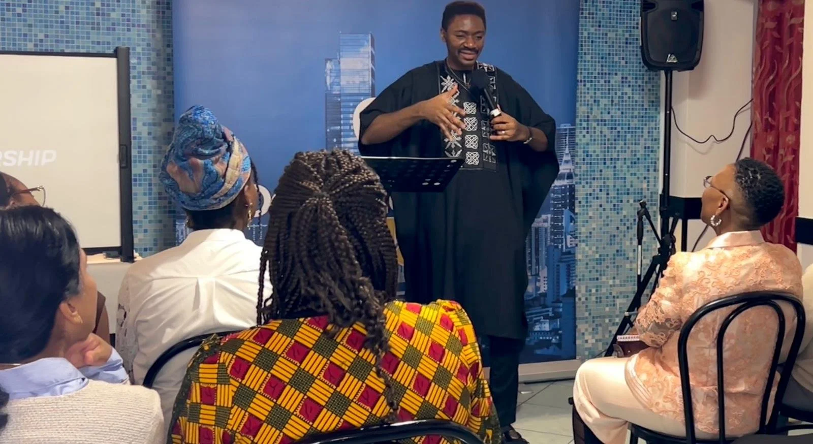 A man in traditional African attire speaks into a microphone during a presentation or seminar, with an audience of women listening attentively in a room with a cityscape backdrop.