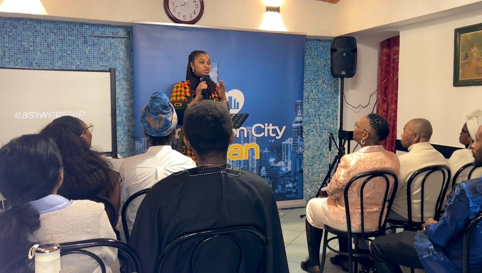 A woman standing at a podium speaking to an audience in a room with a blue backdrop that reads "Urban City" and features skyscraper graphics. The audience is focused on the speaker, with diverse individuals seated in chairs, some wearing glasses, sca