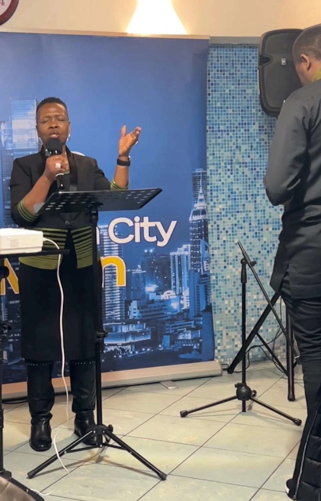 A woman singing into a microphone while standing at a stand with a cityscape backdrop that says 'City'. She is wearing black attire with green striped accents on sleeves, with short hair. Another person, whose face is not visible, stands to the right