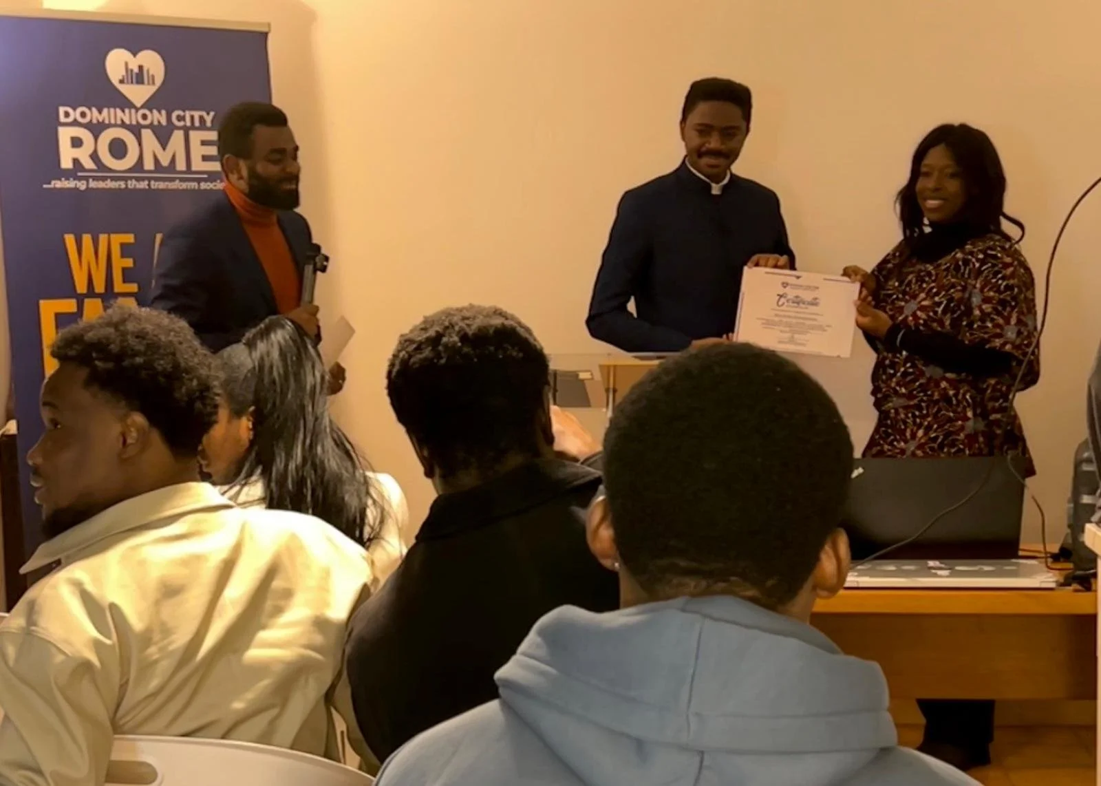 A woman in a patterned dress receives a certificate from a man in a suit during a formal event, with a man holding a microphone standing nearby, and an audience of people watching.